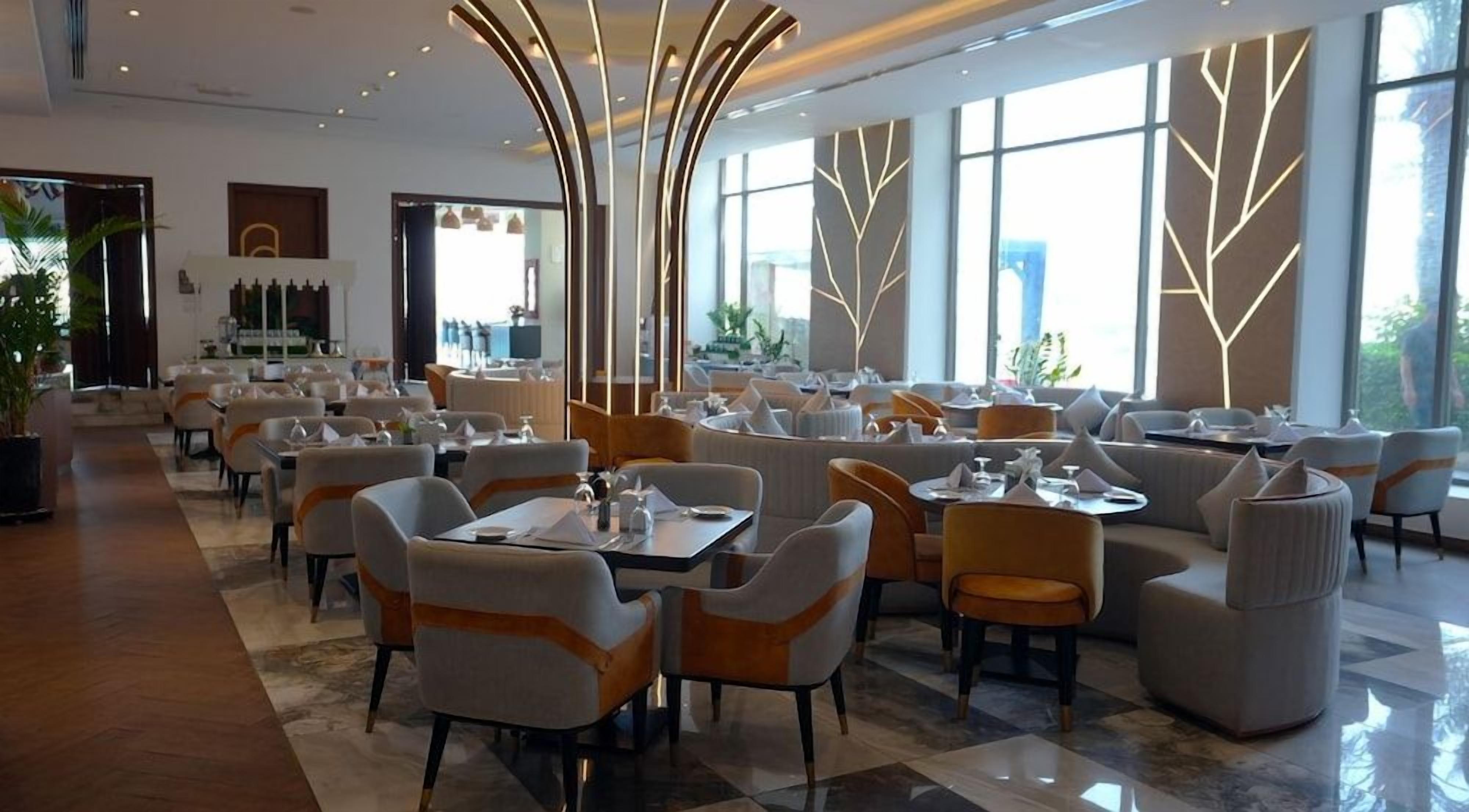 Un amplio comedor de hotel con diversas disposiciones de asientos, incluyendo mesas y sillas, y grandes ventanales que ofrecen luz natural.