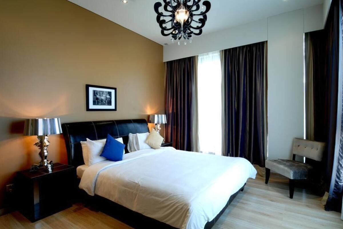 Ein Hotelzimmer mit einem großen Bett, einem Nachttisch, einem Sessel, einer Chandelüster und einem fenster mit Vorhängen.
