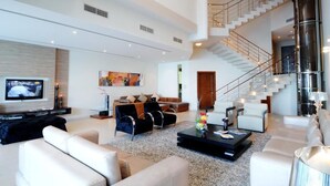A spacious living area with a large flat-screen TV, a modern staircase, and a high ceiling. - The Dragon Beach Hotel & Resort (Amwaj Islands)