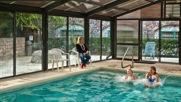 An indoor swimming pool with clear glass walls, a diving board, and a few chairs around the pool. - Helendorf River Inn, Suites & Conference Center (Helen)