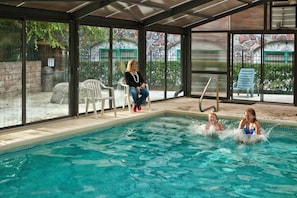 Piscina interior com paredes de vidro transparente, plataforma de salto e algumas cadeiras ao redor.