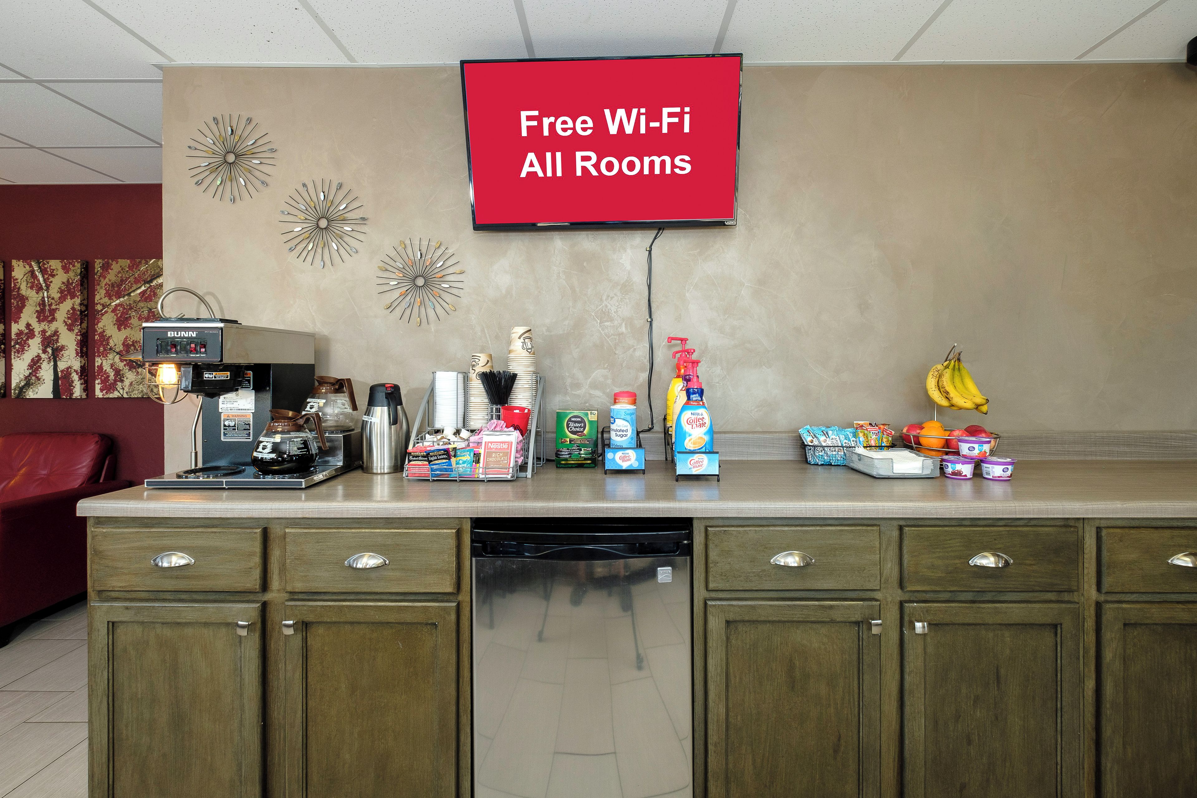 A hotel reception area with a coffee station, a flat-screen TV displaying 'Free Wi-Fi All Rooms,' and various food and drink items.