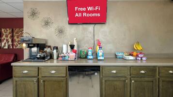 A hotel reception area with a coffee station, a flat-screen TV displaying 'Free Wi-Fi All Rooms,' and various food and drink items.