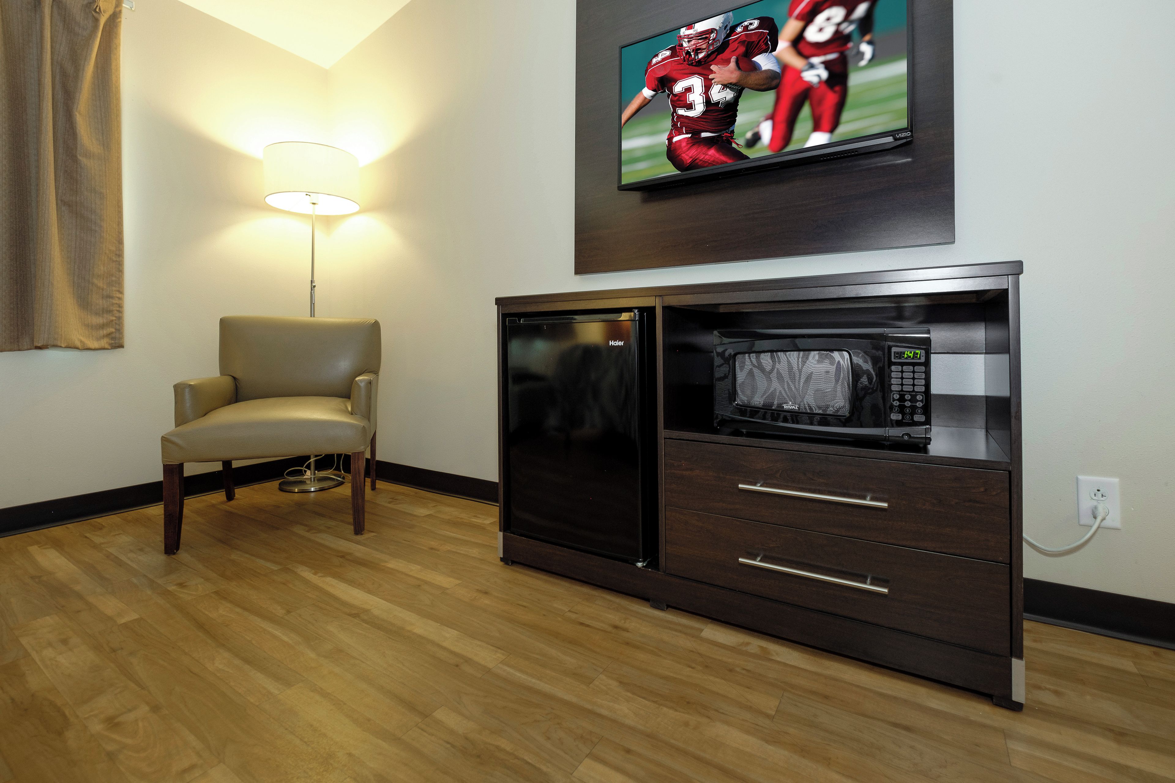 Habitación de hotel con televisor de pantalla plana montado en la pared, una silla moderna, microondas integrado y piso de madera.