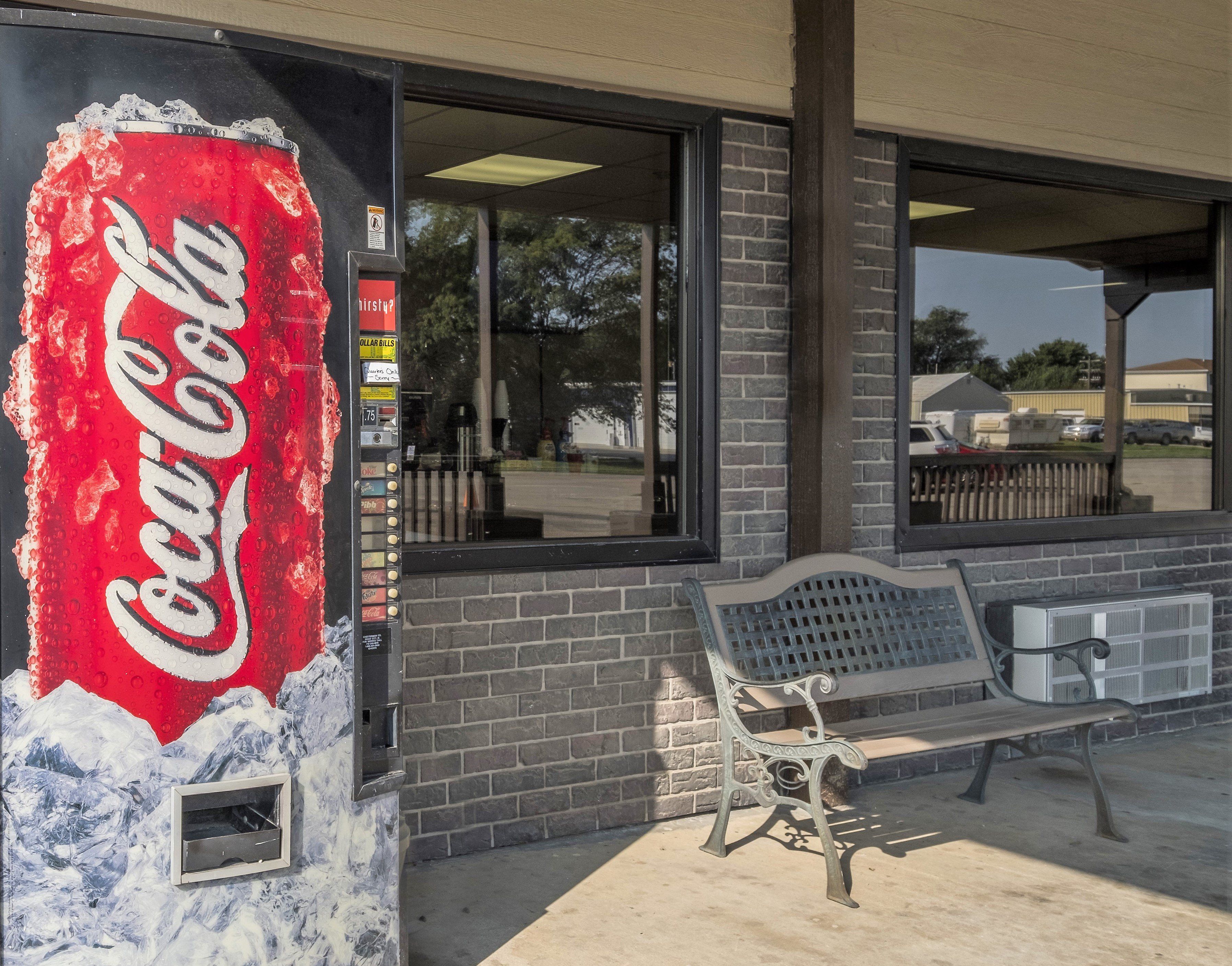 Una máquina expendedora de Coca-Cola frente a un edificio de fachada de ladrillo y una banca.
