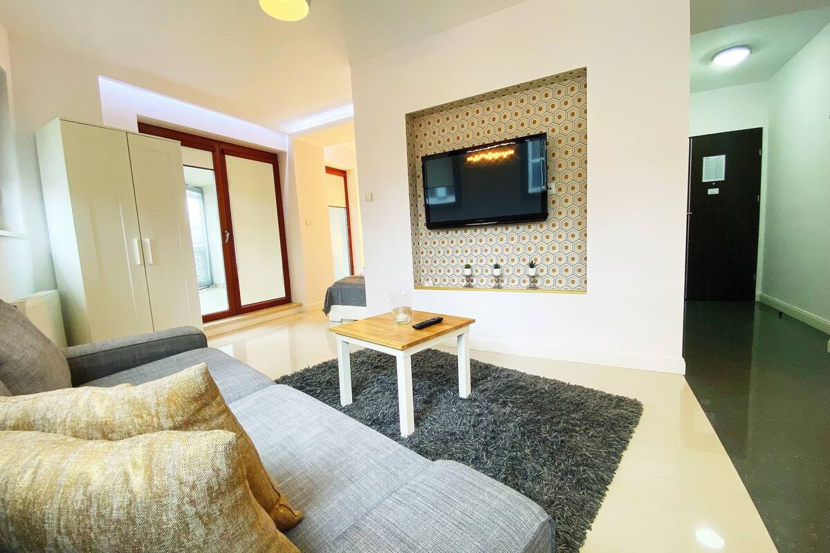 A modern living room with a grey sofa, a small wooden table, and a TV mounted on a patterned wall.