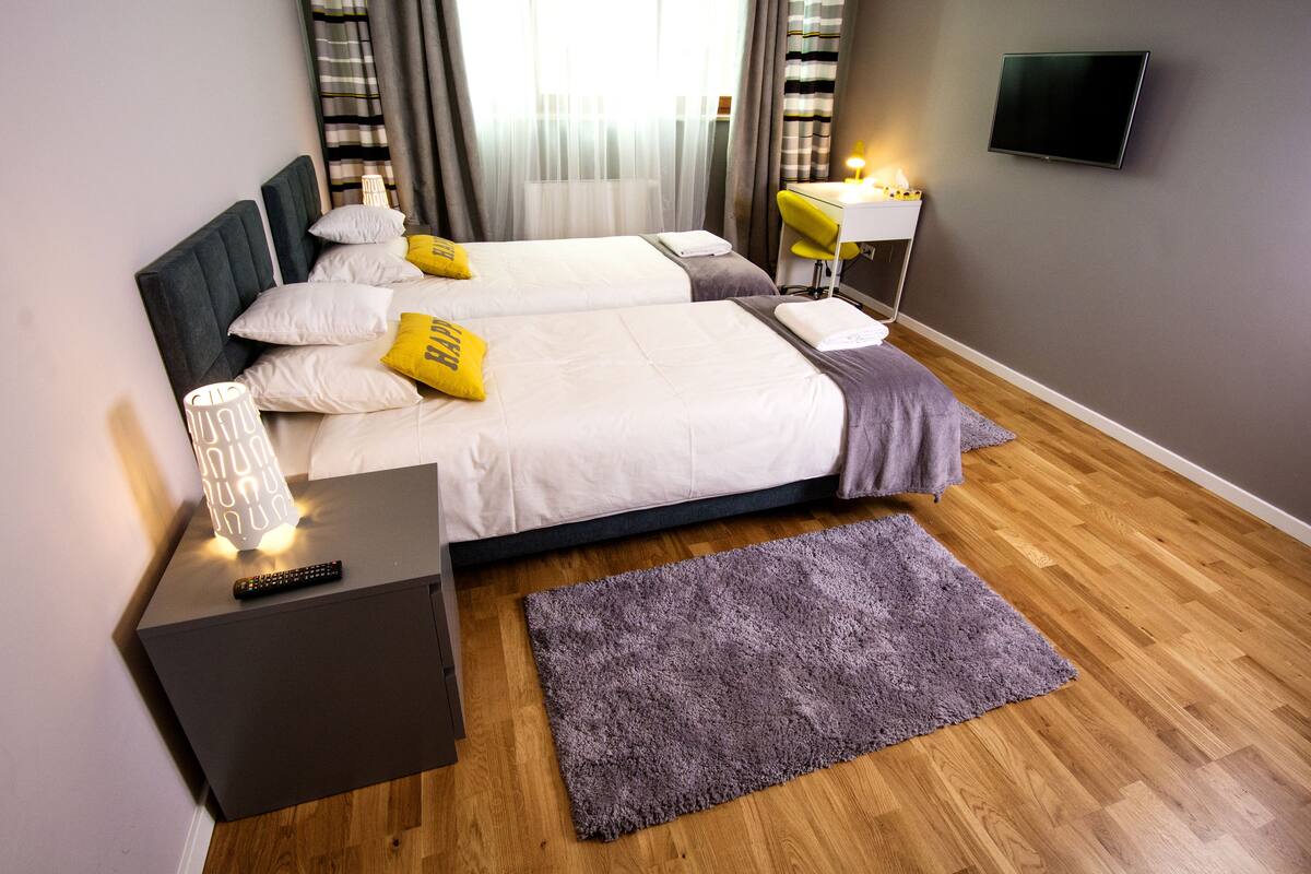 A double bed with white and yellow pillows, a bedside table with a lamp, a TV mounted on the wall, and a wooden floor.