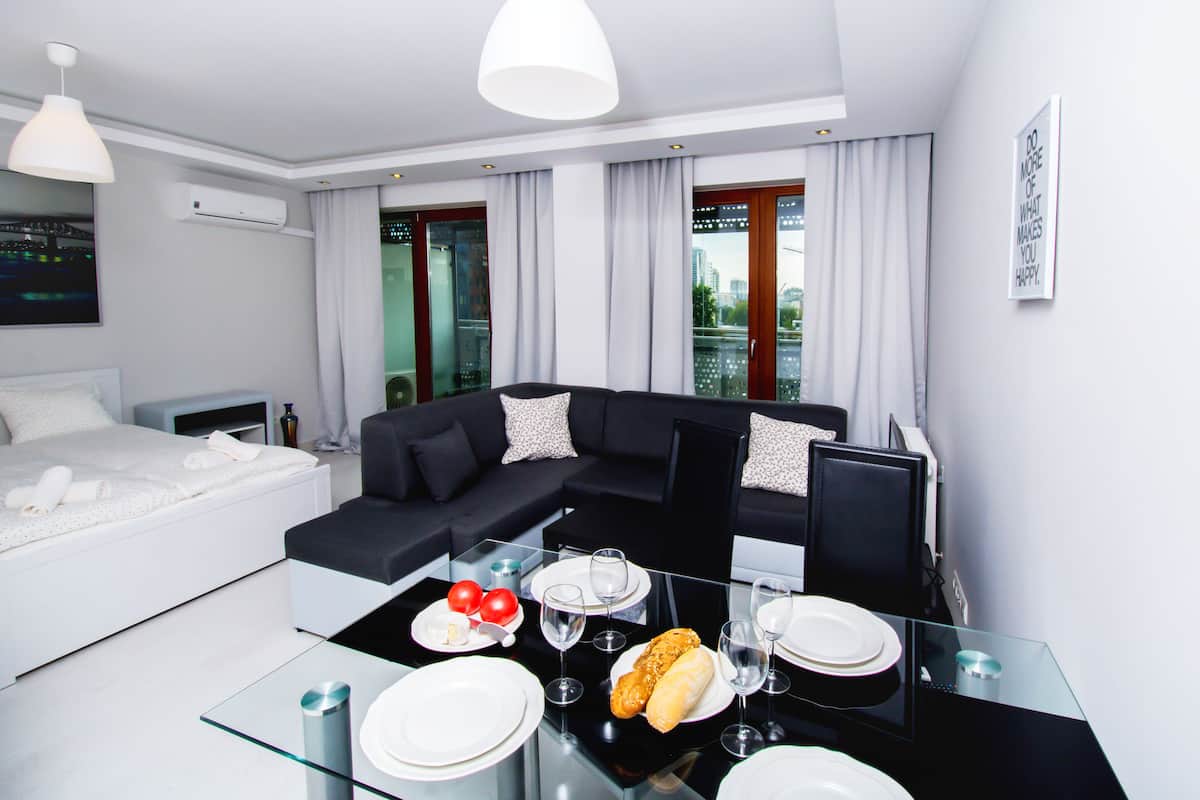 A modern hotel room with a bed, a black sectional sofa, a glass dining table set with plates, glasses, and a baguette.
