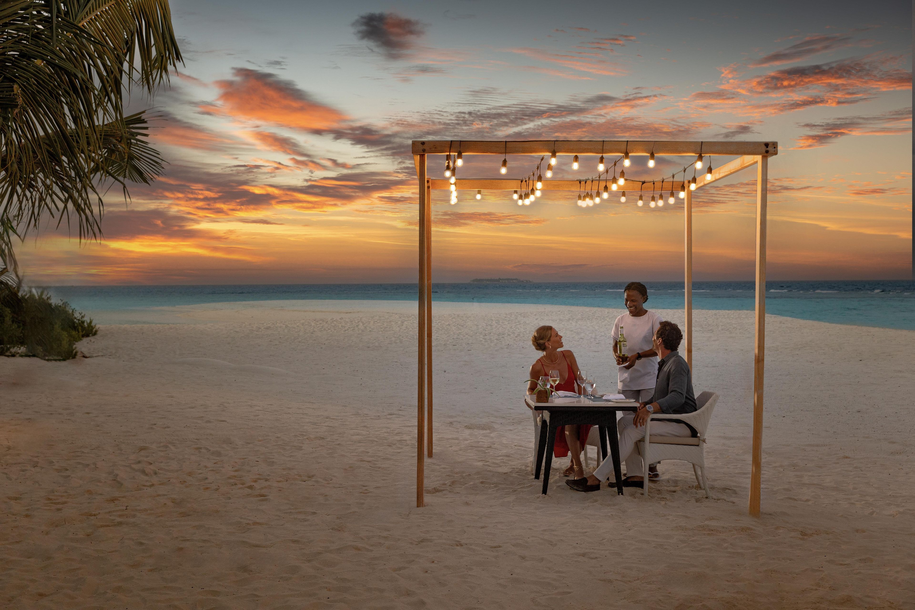 A beachside dining setup with a table and chairs under a wooden pergola with string lights.