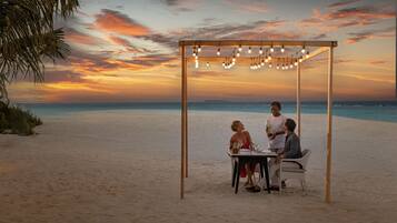 A beachside dining setup with a table and chairs under a wooden pergola with string lights.