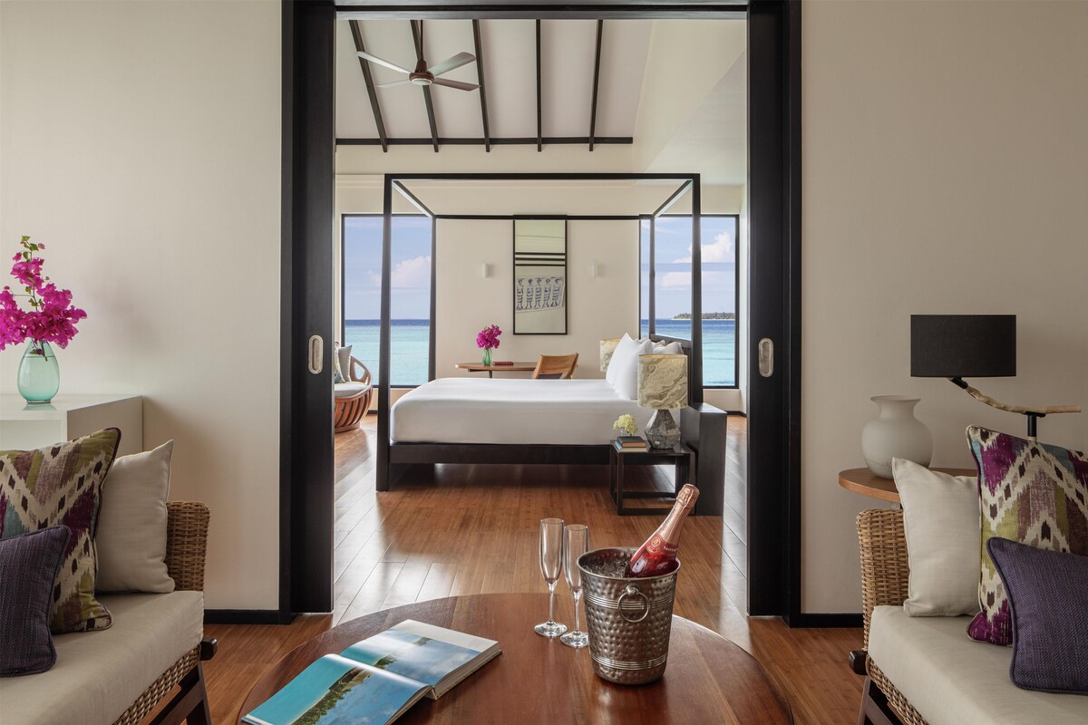 A hotel room with a bed, a view of the sea, a balcony, and a dining area with a bottle of champagne.