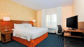 A hotel room with a large bed, a desk with a TV, a chair, a desk lamp, a bedside table, and a window with curtains.