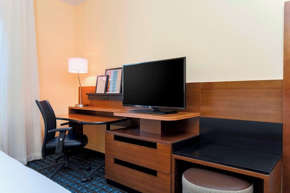 A hotel room with a wooden desk, a television, a chair, a lamp, and a small table.