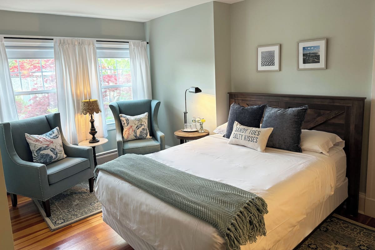 Quarto com cama grande, duas poltronas, mesa de cabeceira e janela com cortinas.