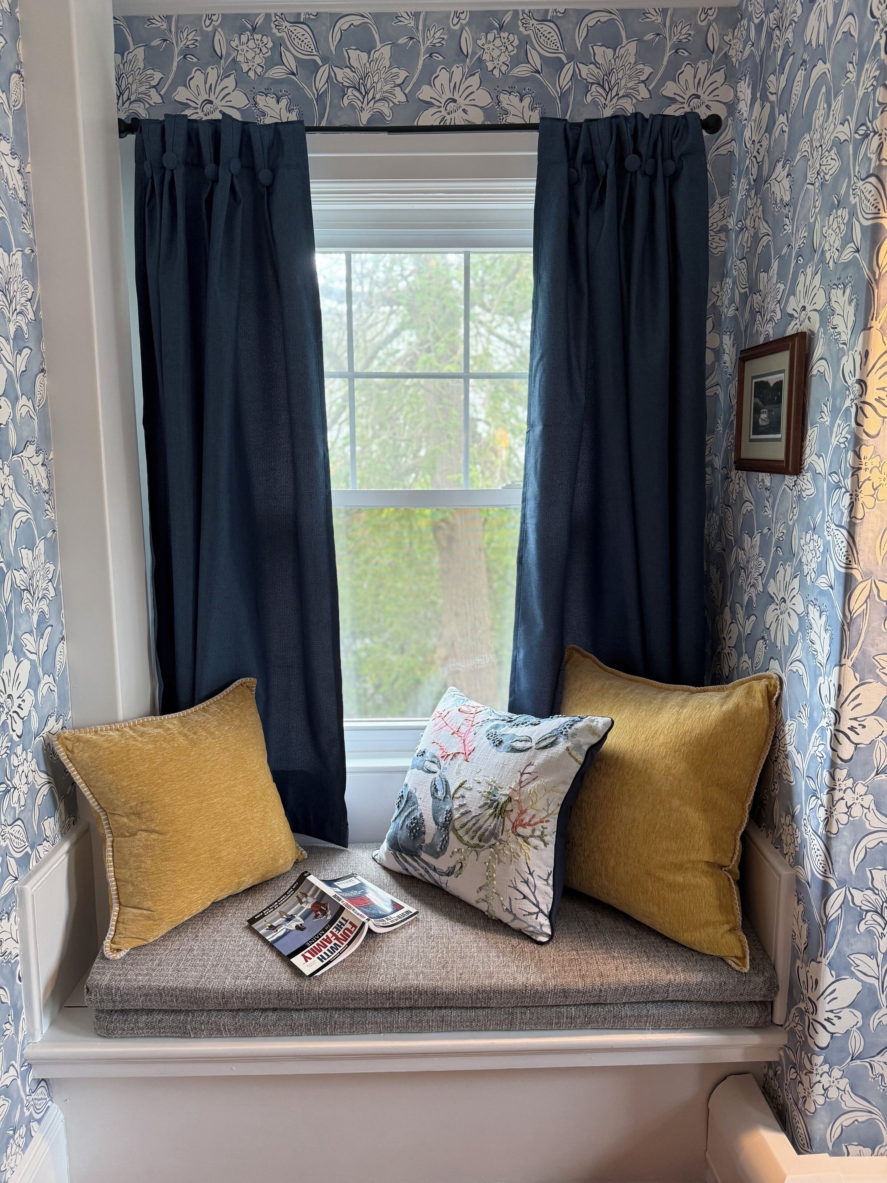 Ein Fensterplatz mit zwei Kissen, einer Zeitschrift und einem gerahmten Bild an der Wand.
