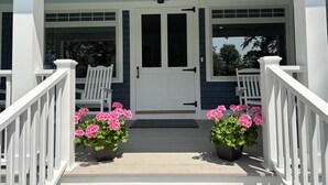 A front porch with a white door, two white chairs, and two potted pink flowers.