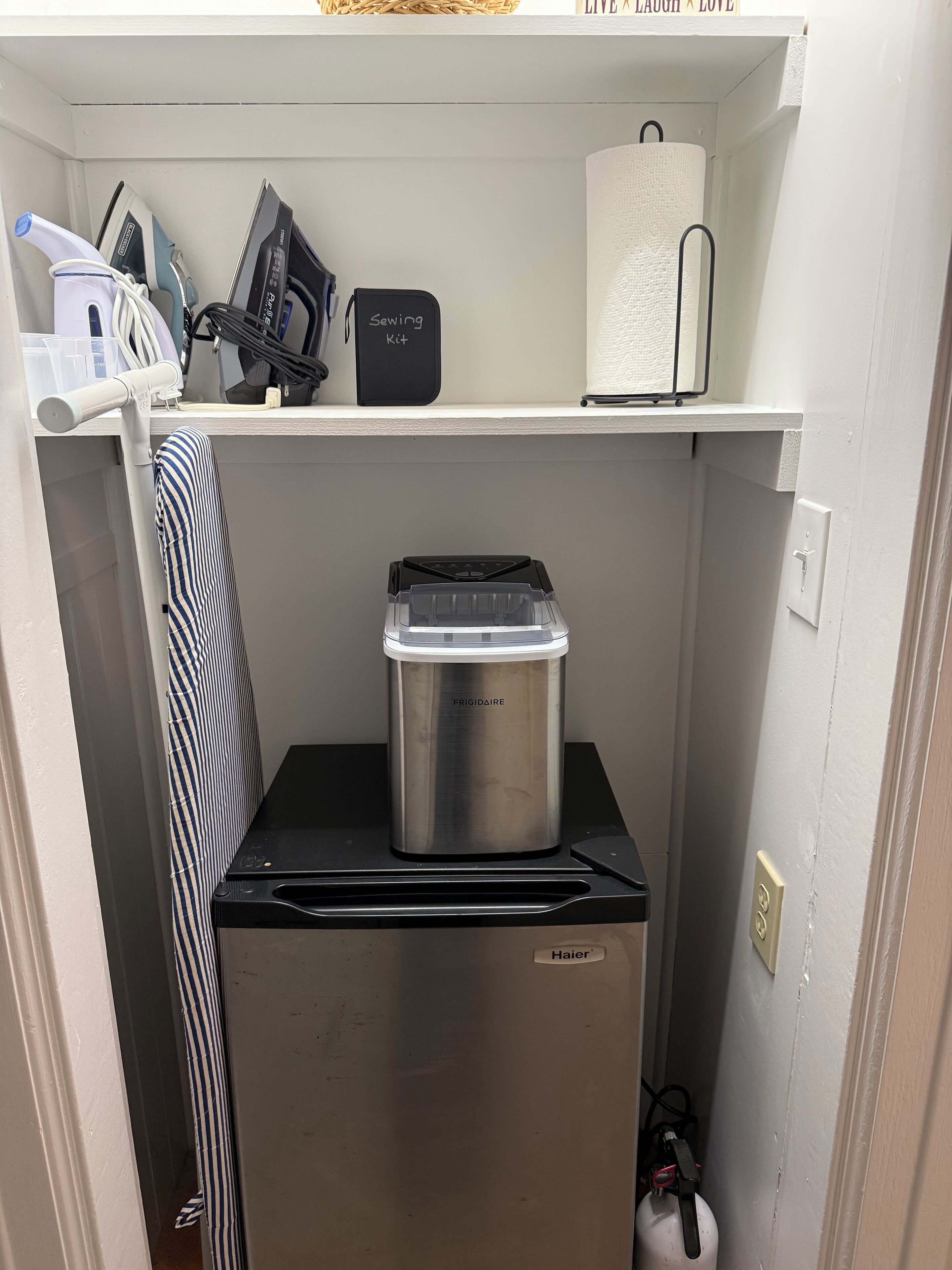 Una kitchenette compacta con un refrigerador de acero inoxidable, un secador de pelo negro y un kit de costura en un estante.