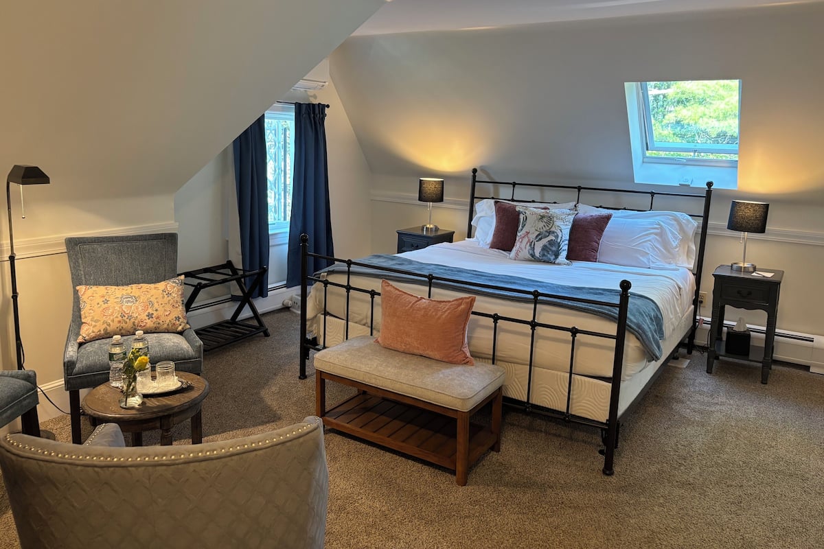 Um quarto com cama, cadeira, mesa pequena e janela.