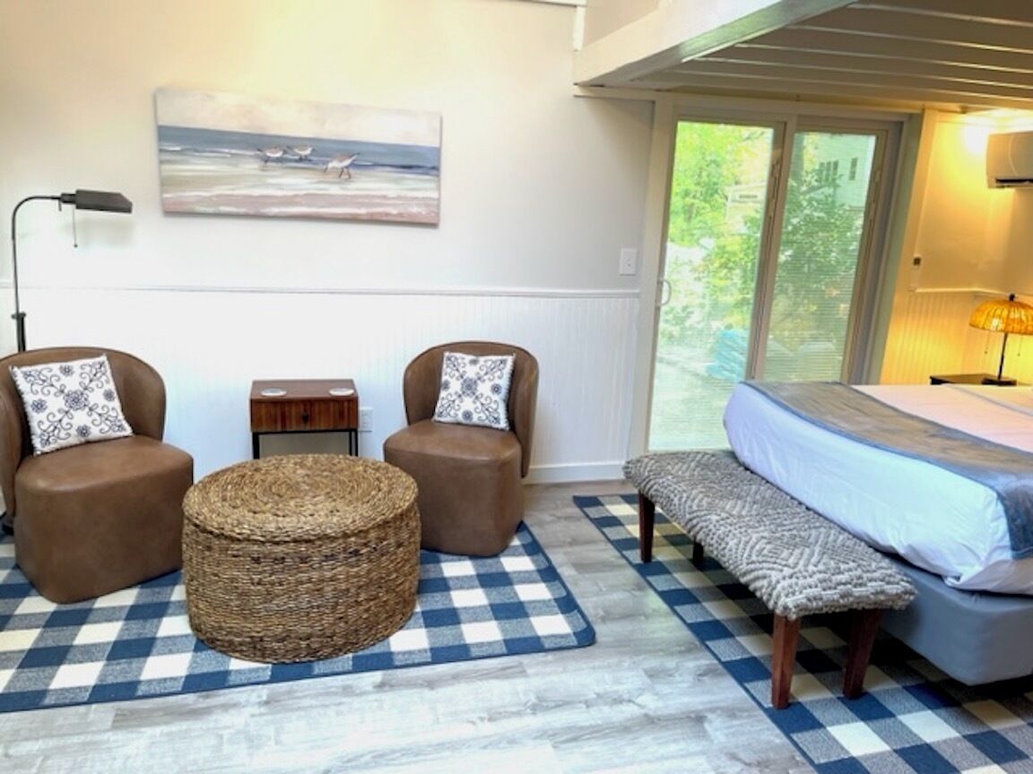 A room with a bed, two armchairs, a round woven ottoman, a bench, a checkered rug, a small wooden side table, and a painting of birds on the wall.