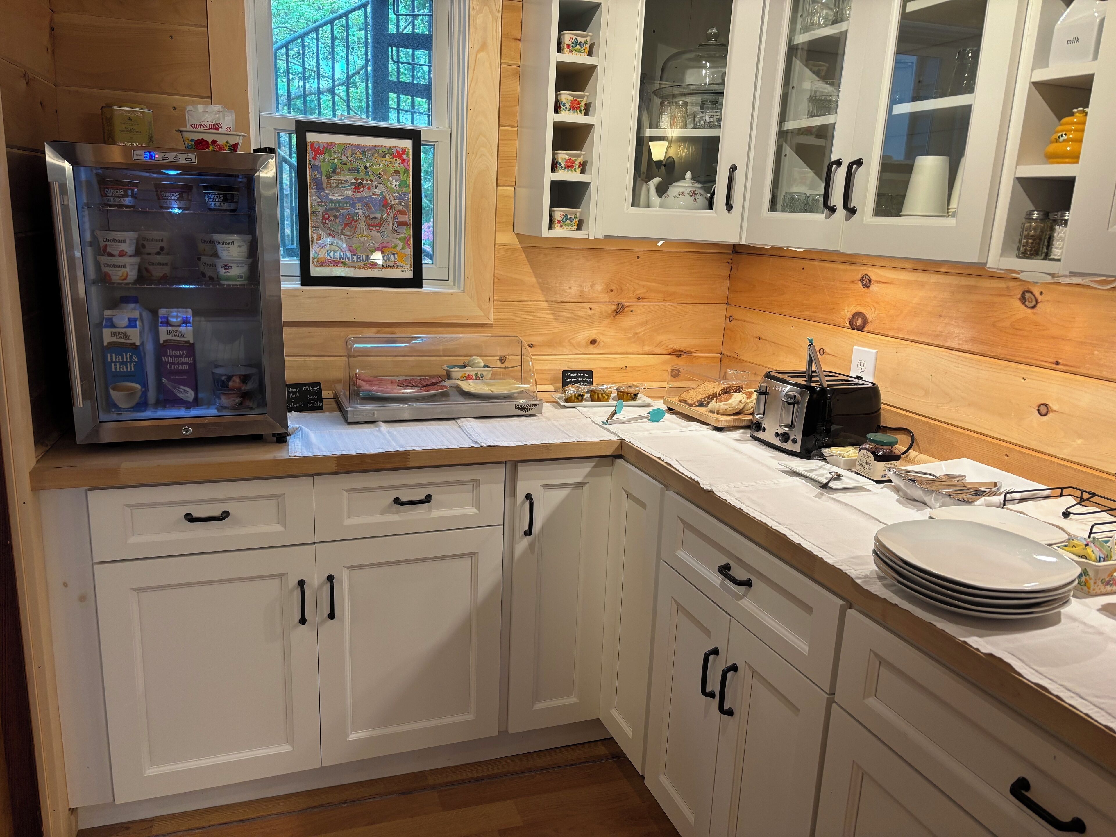 Une cuisine avec des meubles blancs, un réfrigérateur, une fenêtre donnant sur un beau paysage et un plan de travail sur lequel sont disposés divers objets.