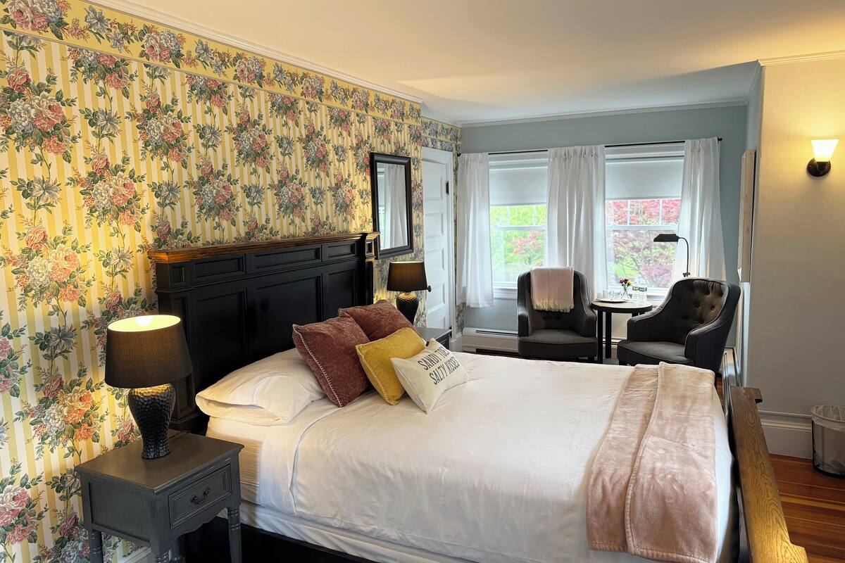 Quarto de hotel com uma cama grande, uma mesa de cabeceira, uma escrivaninha com duas cadeiras e uma janela com cortinas.