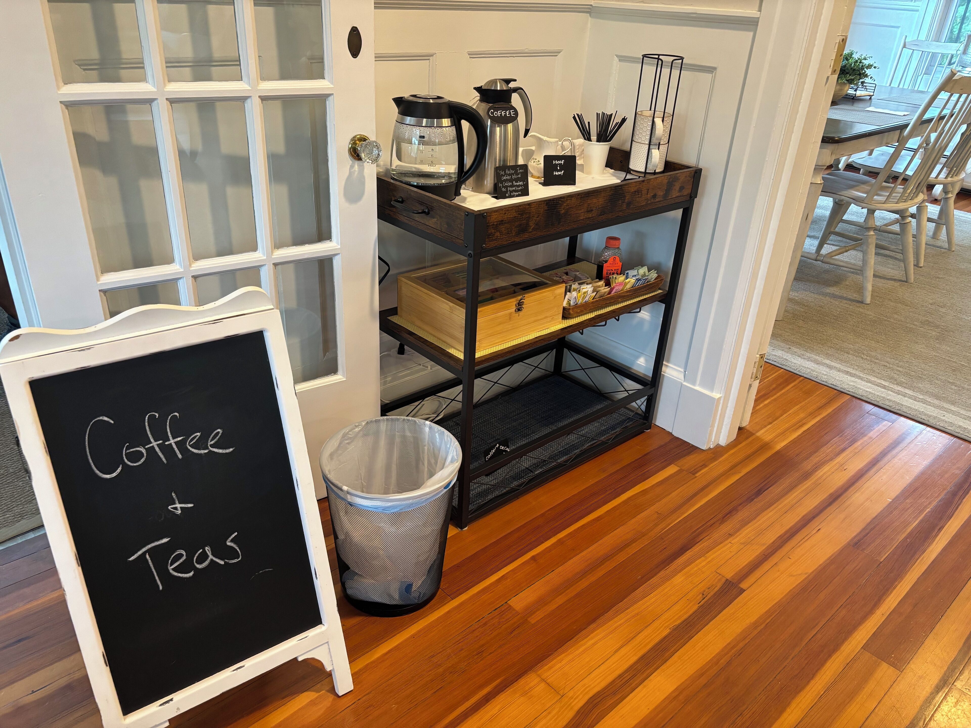 A coffee and tea station with a chalkboard sign, a coffee maker, and a trash can.