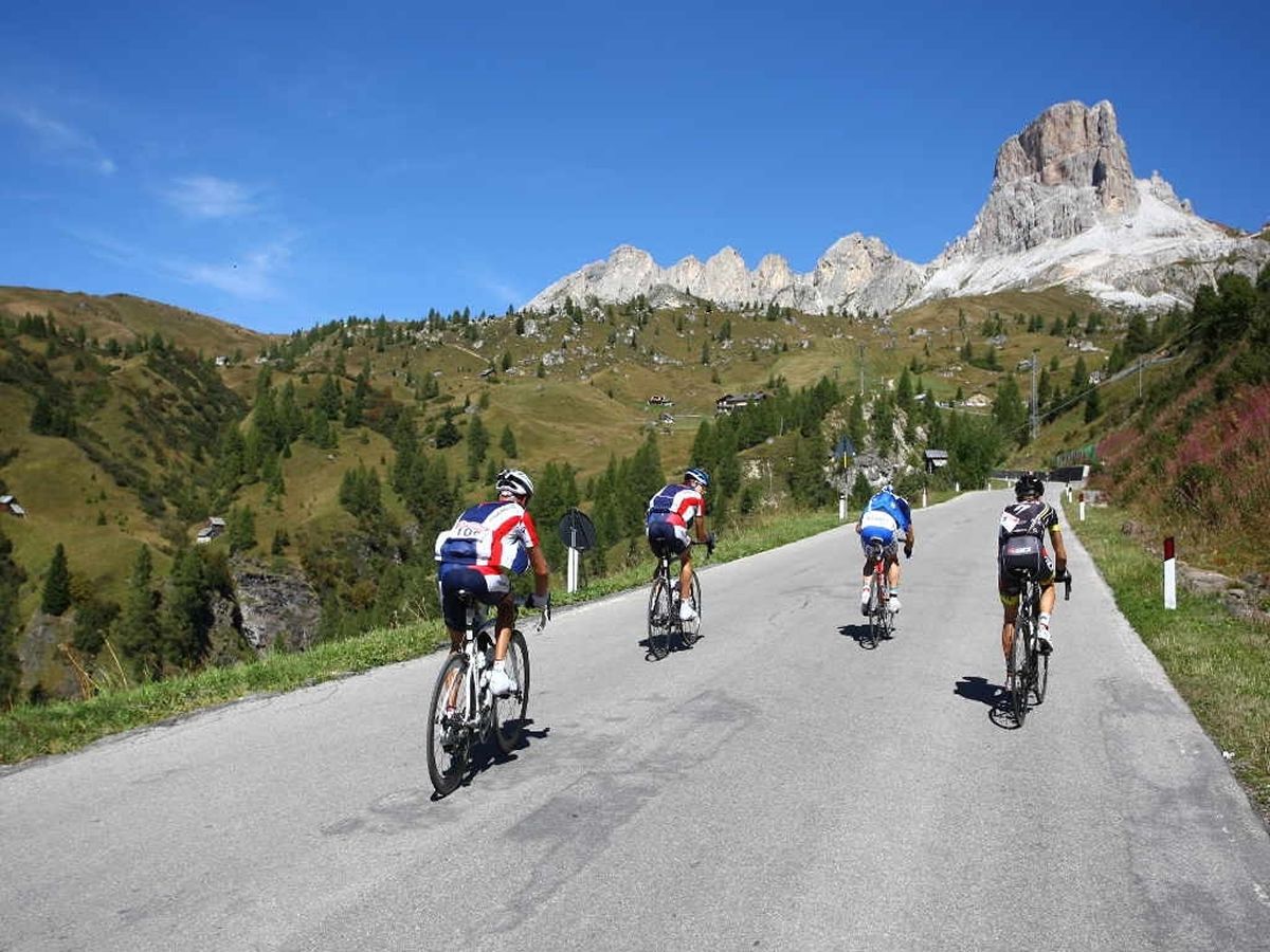 A paved road with cyclists in motion, surrounded by green hills and a prominent mountain peak in the background.