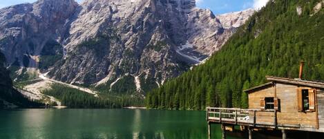 A wooden cabin on stilts by a clear green lake with a mountain backdrop.
