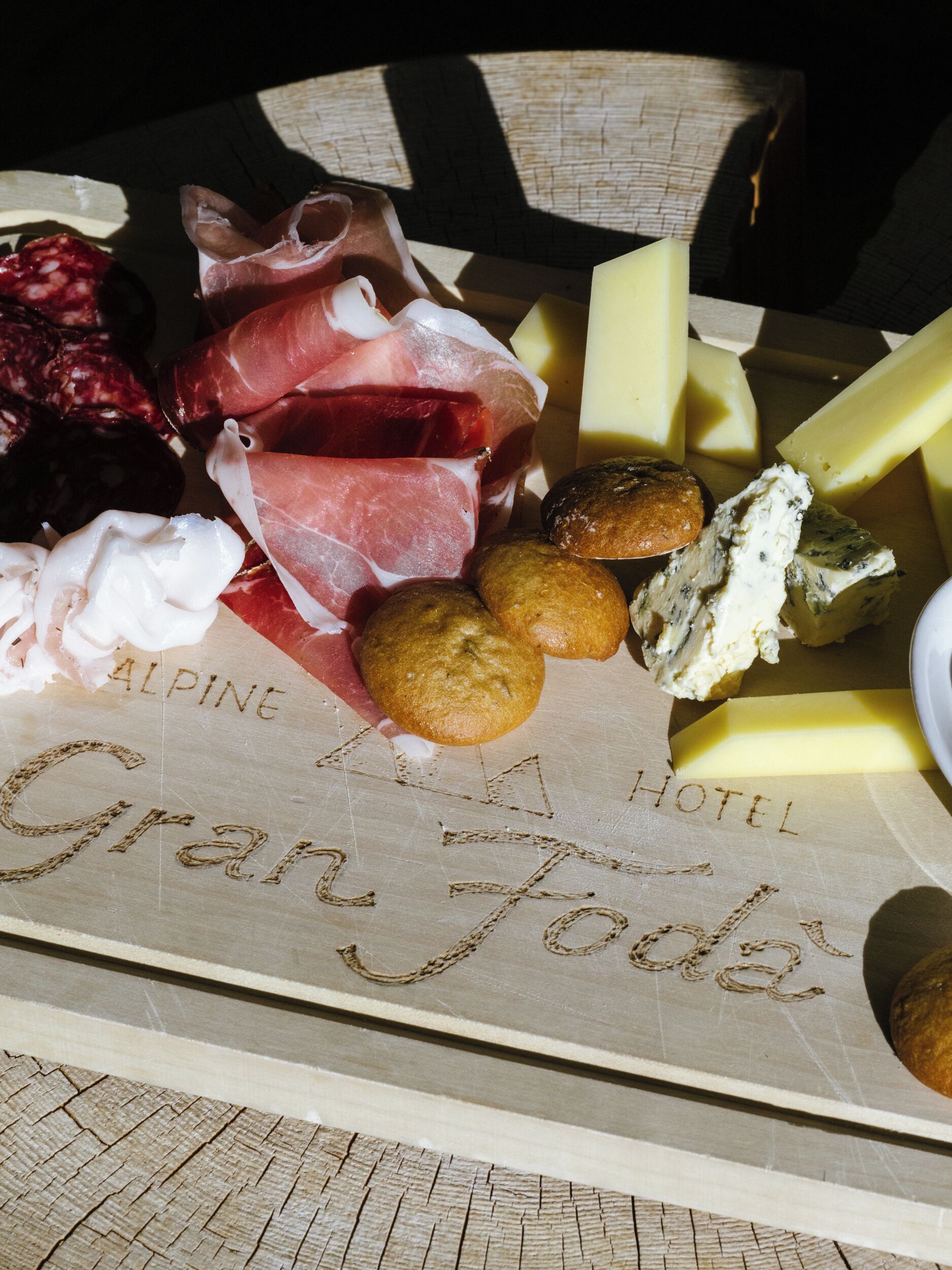 A charcuterie board with various meats, cheeses, and bread on a wooden surface with the text "Alpine Hotel Gran Jode" engraved on it.