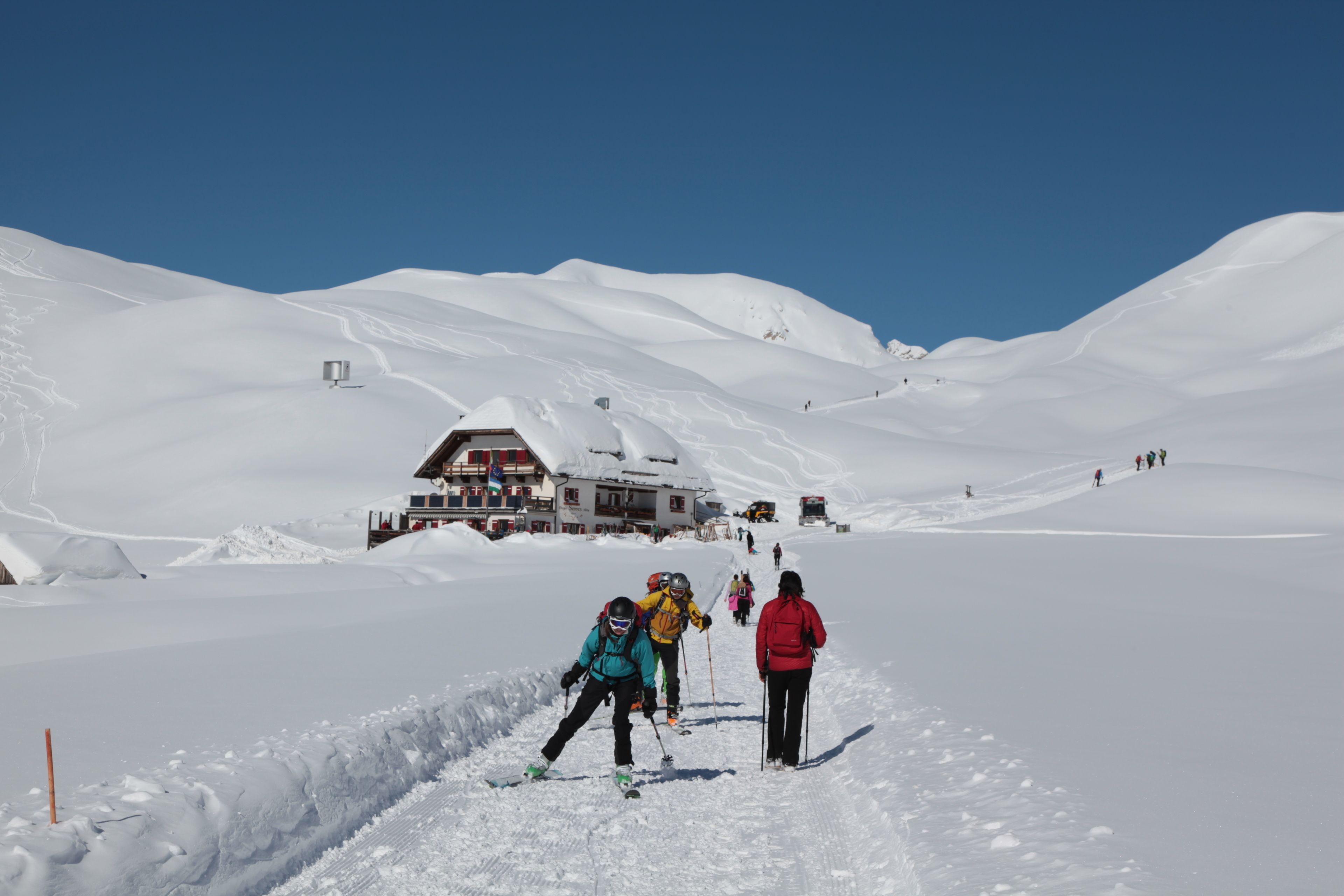 A snow-covered ski slope with skiers, a lodge, and a clear blue sky.