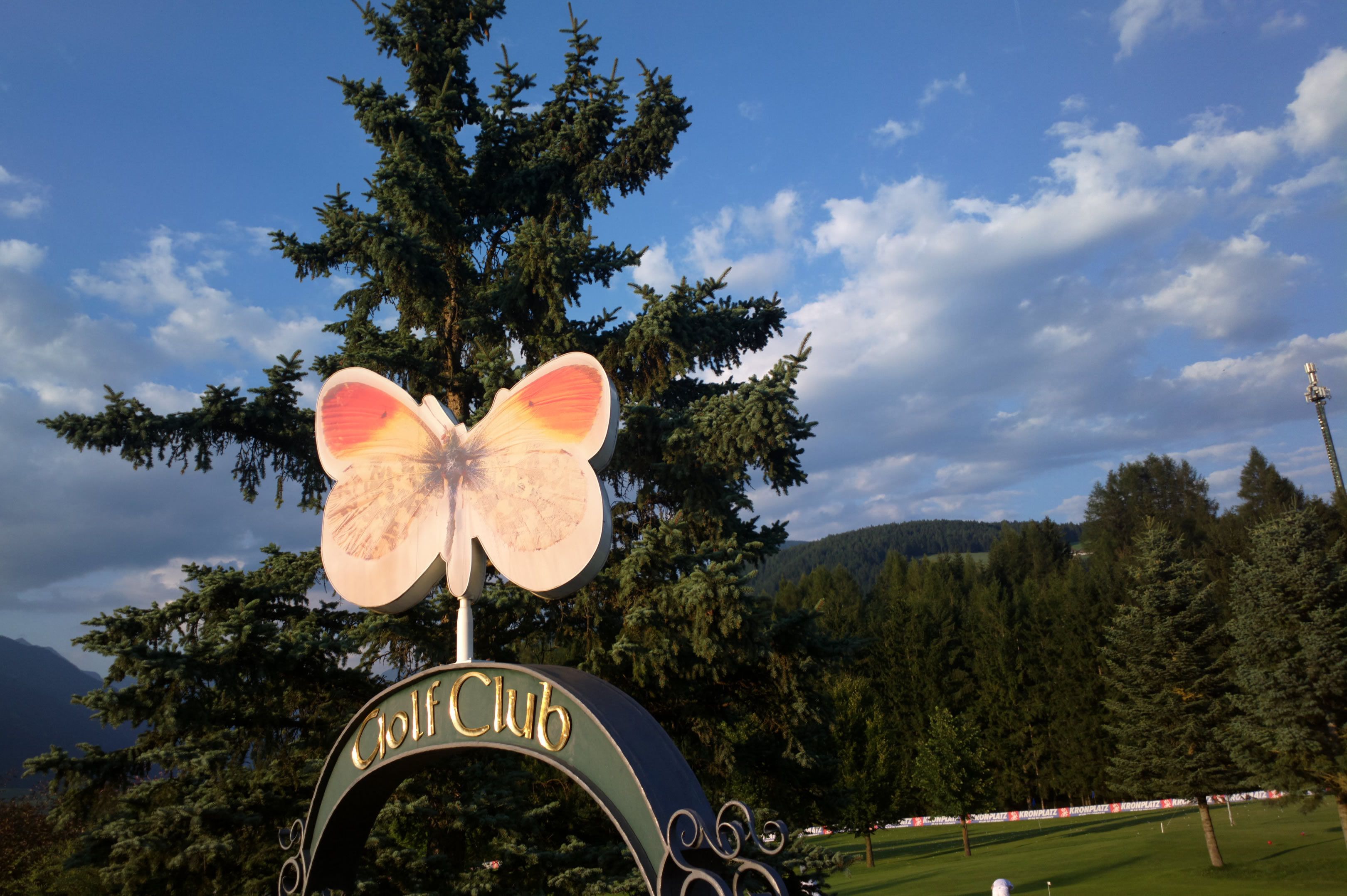 A golf club sign with a butterfly design, surrounded by trees and a mountainous backdrop.