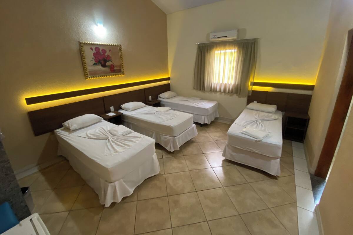 A hotel room with four beds arranged in a U-shape, a painting on the wall, an air conditioning unit, and a window with curtains.