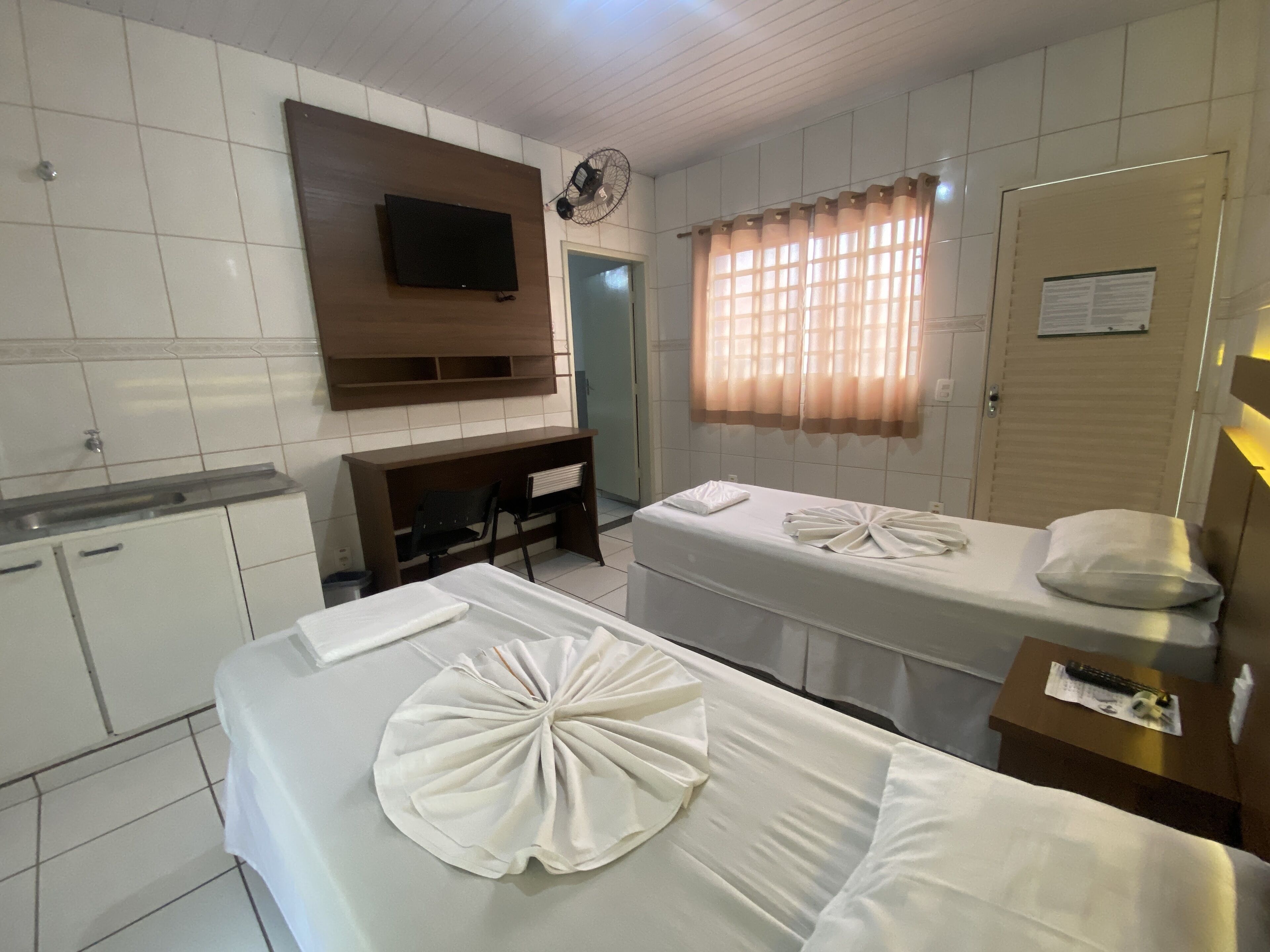 Habitación de hotel con dos camas, un escritorio, un televisor, un ventilador y una ventana con cortinas.