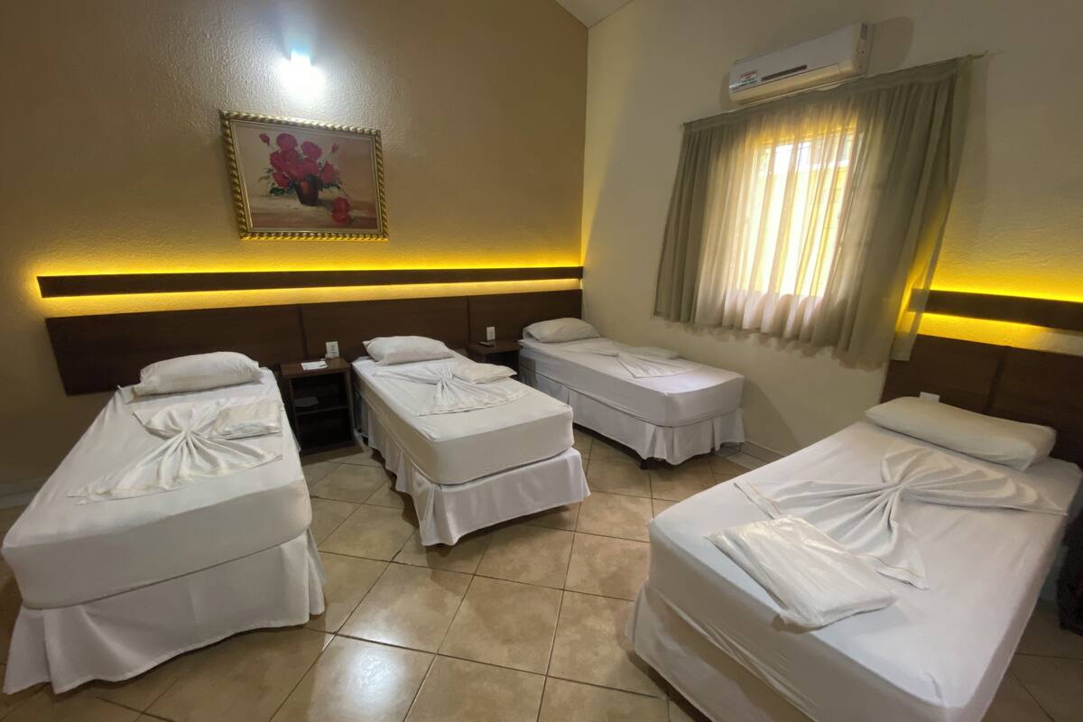 A hotel room with four single beds, a bedside table with a lamp, a wall-mounted air conditioner, and a framed picture above the beds.