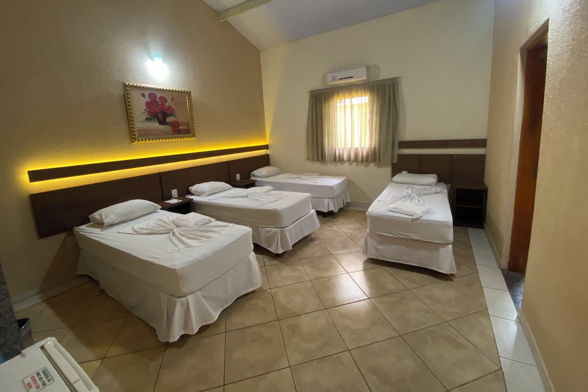Quarto de hotel com quatro camas, ar condicionado fixado na parede, um quadro e uma luminária no teto.