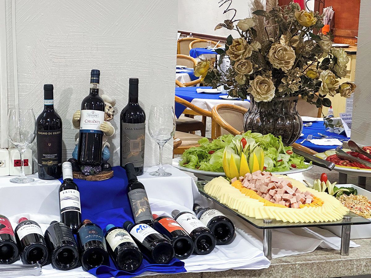 A table with multiple bottles of wine, a vase with artificial flowers, and a variety of food items.