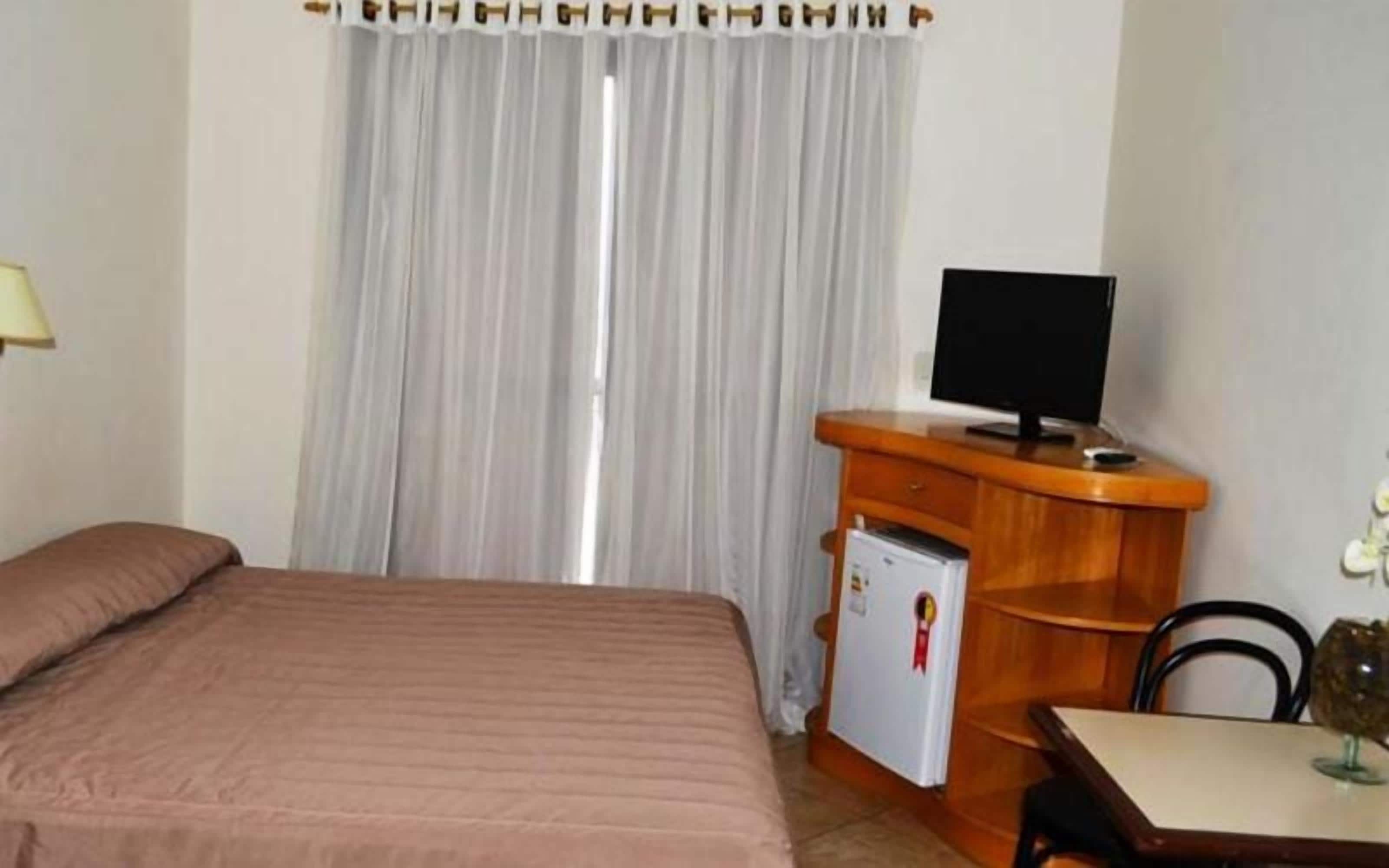 Ein Hotelzimmer mit einem Bett, einem Schreibtisch mit Computer, einem kleinen Kühlschrank und einem Fenster mit Vorhängen.