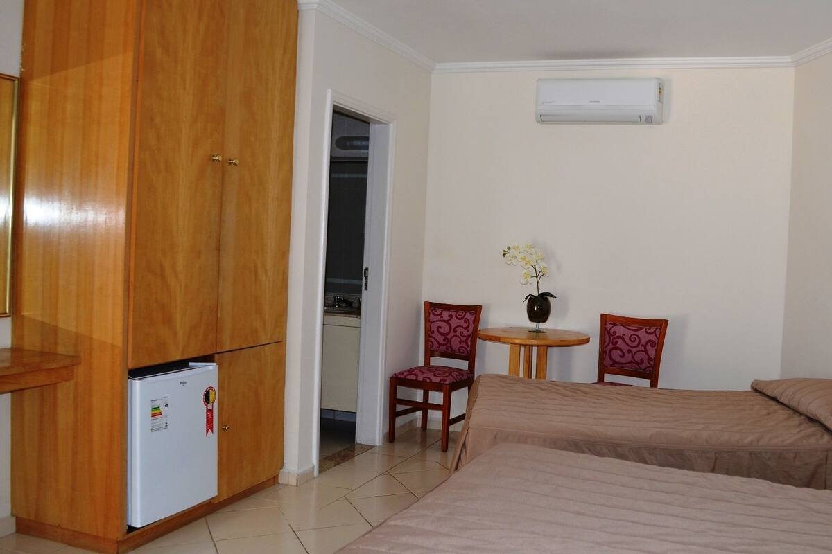 Ein Hotelzimmer mit Holzschrank, kleinem Kühlschrank, zwei Betten, Tisch mit Vase und Wandklimaanlage.