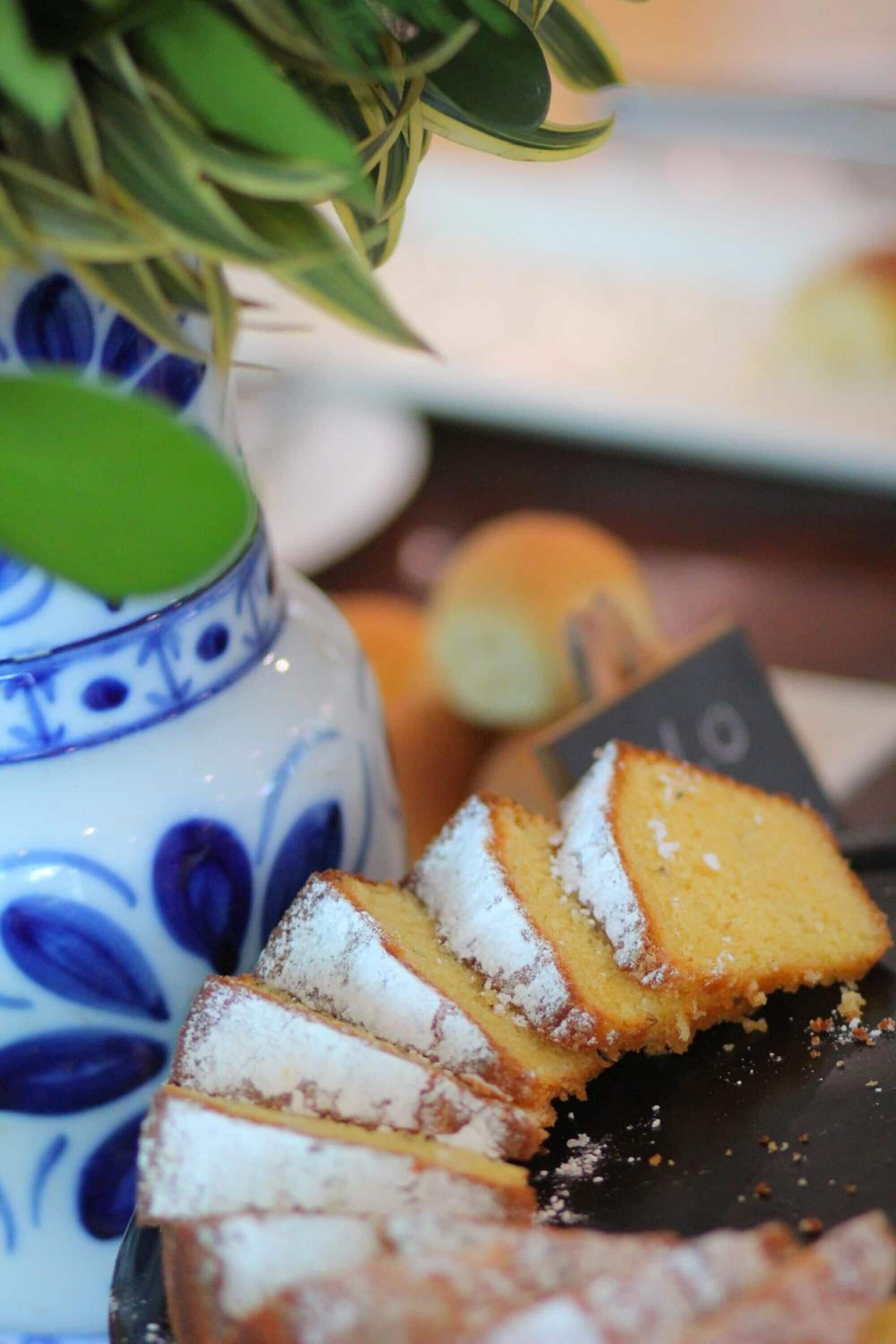 A close-up of a cake with powdered sugar on a plate, a blue and white ceramic vase, and a green plant in the background.