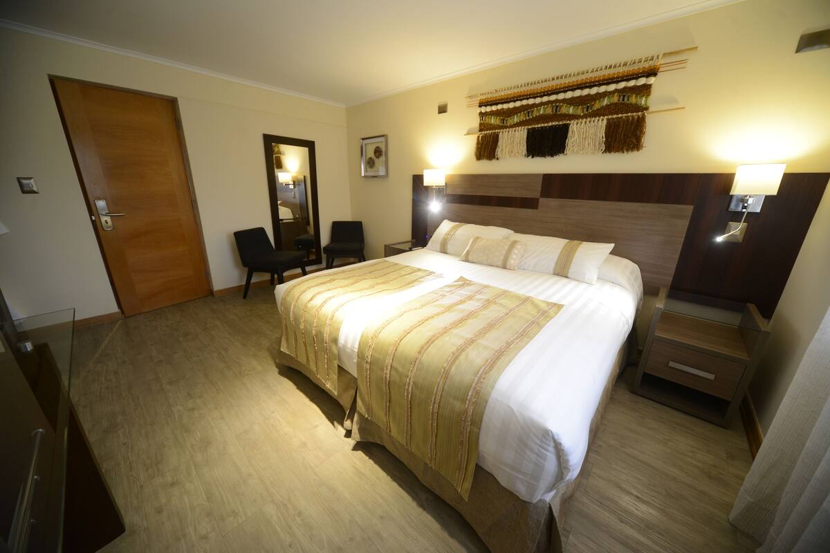 A hotel room with a large bed, a wooden headboard, a bedside table, a chair, a desk, and a mirror.
