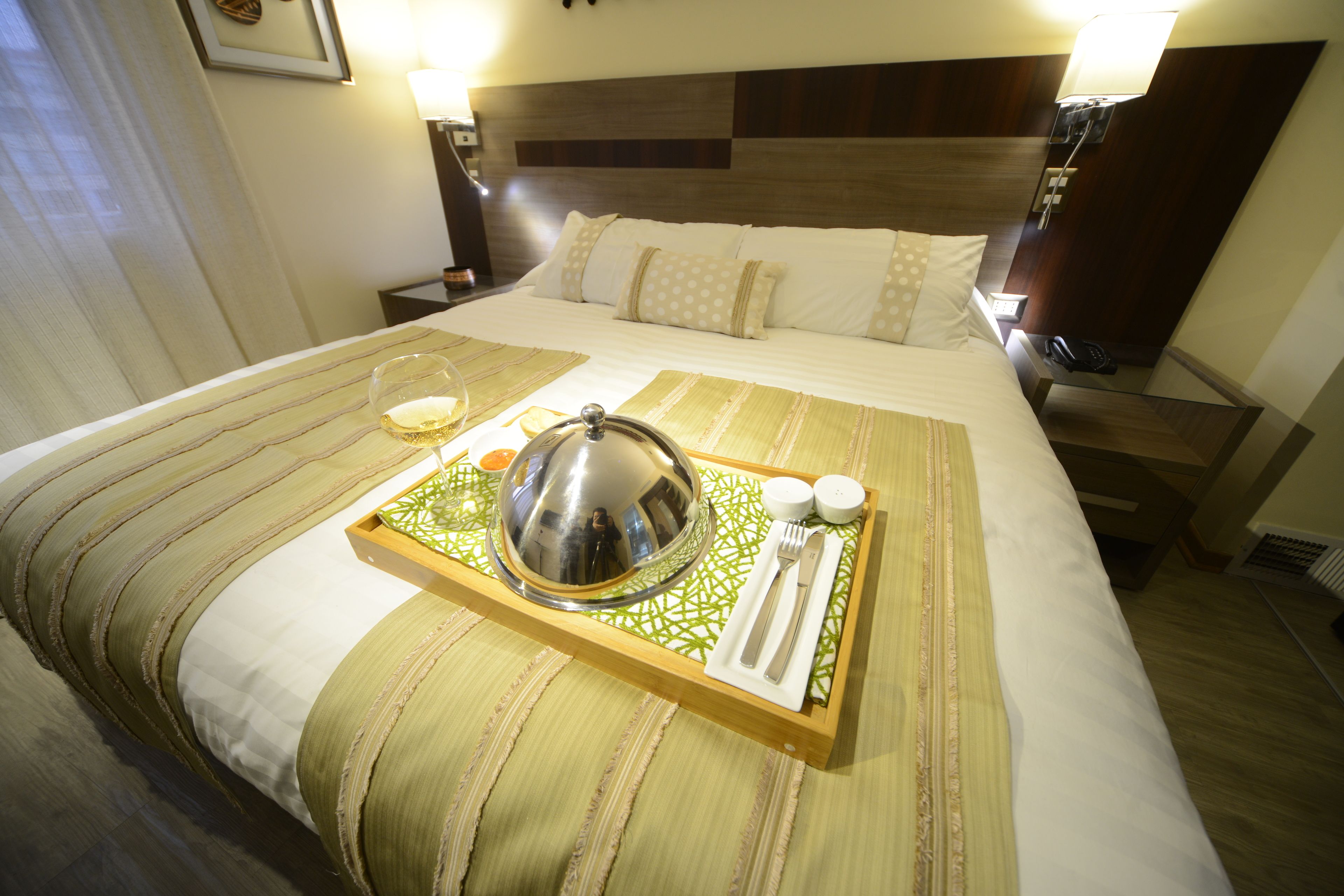 A neatly made bed with a tray containing a glass of wine, a silver bowl, and a set of cutlery.