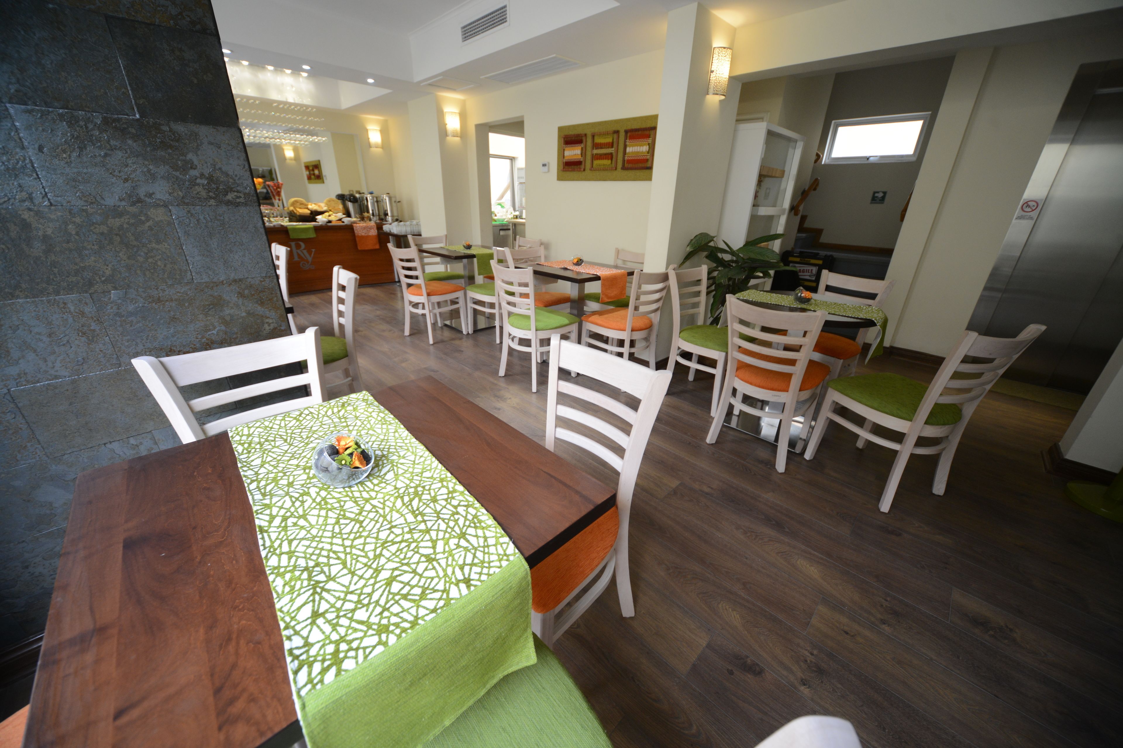 A dining area with multiple tables and chairs, a kitchen in the background, and a stone wall.