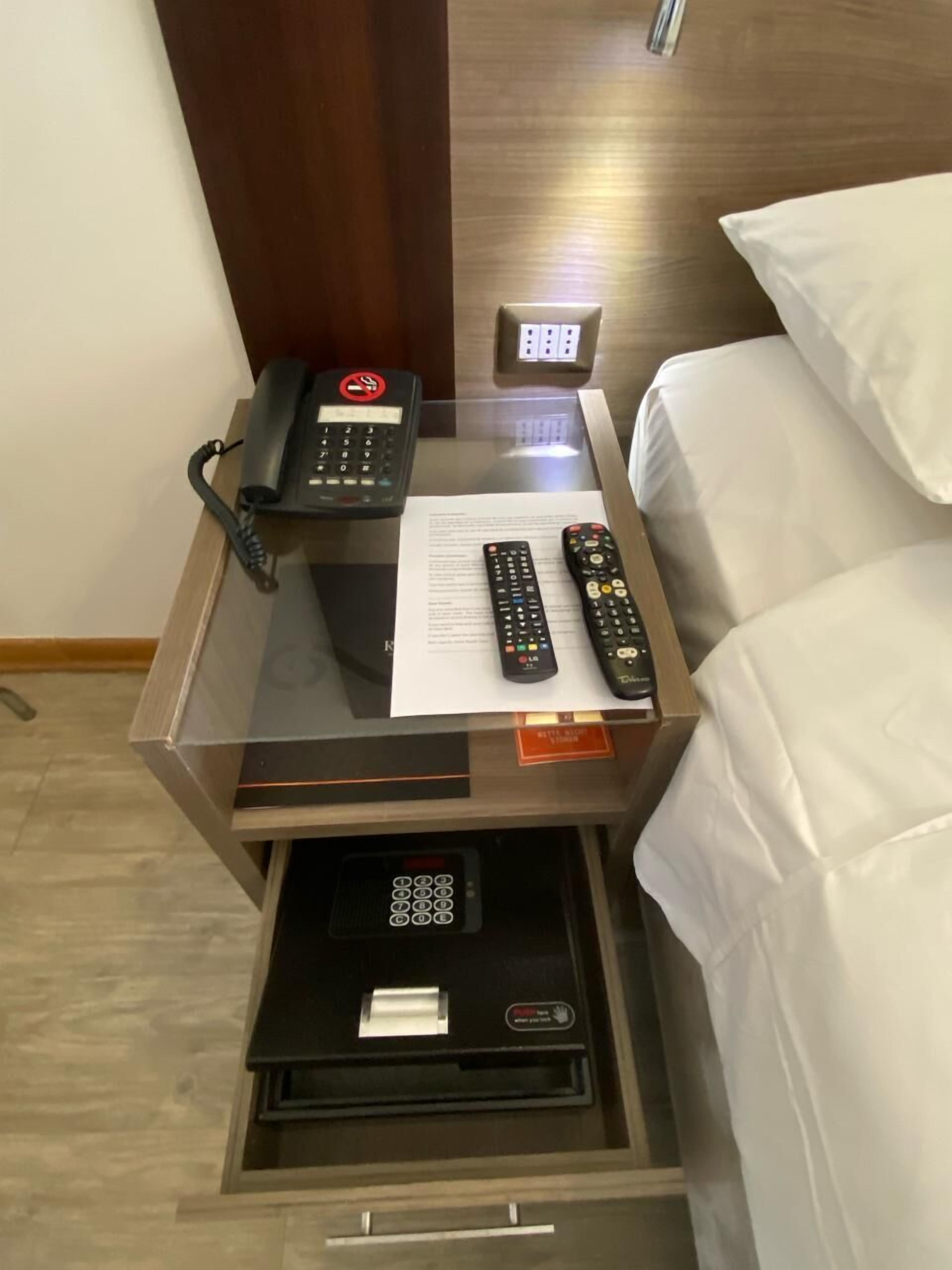A hotel room with a glass-top desk, a telephone, two remote controls, a notepad, and a drawer with a keypad.