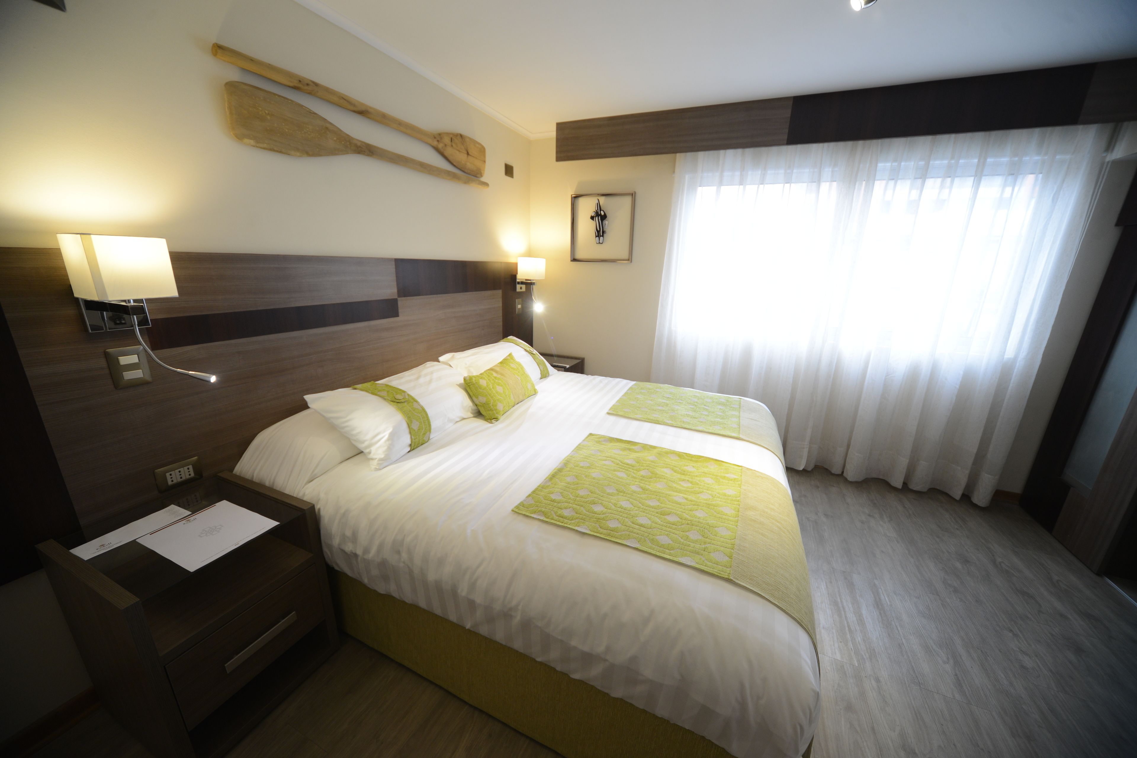 Habitación de hotel con una cama grande, dos mesitas de noche, una lámpara de pared, un cuadro y una ventana con cortinas.