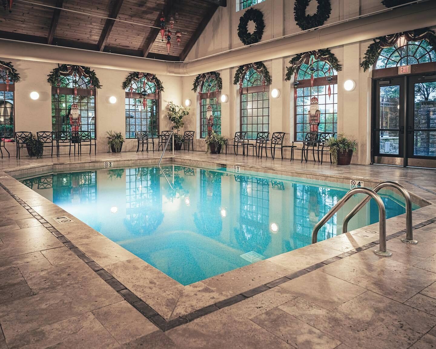 An indoor swimming pool with a tiled floor, surrounded by chairs and potted plants, and decorated with wreaths and Christmas ornaments.