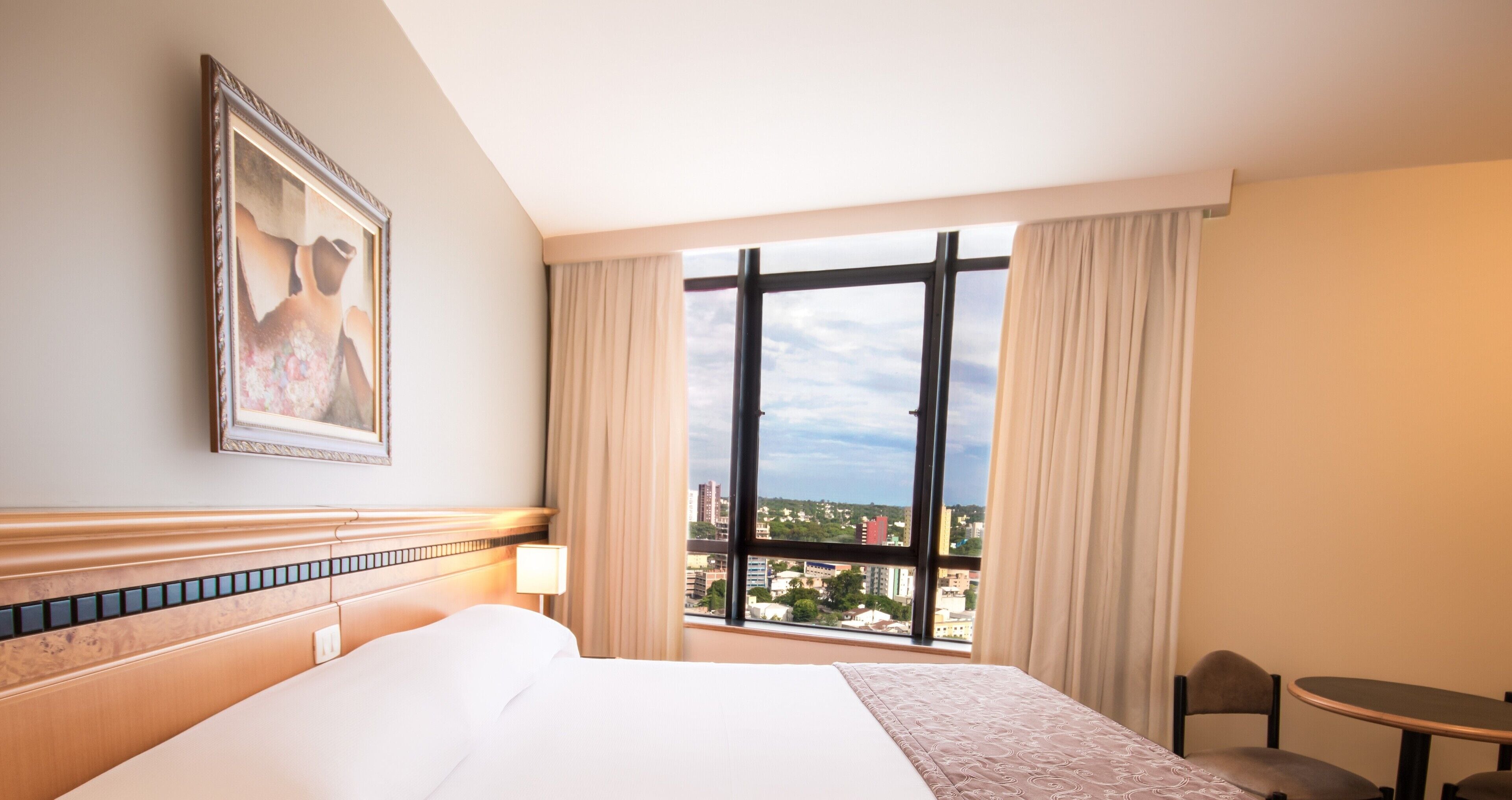 Quarto de hotel com uma cama grande, uma janela com cortinas, uma mesa pequena e um quadro na parede.