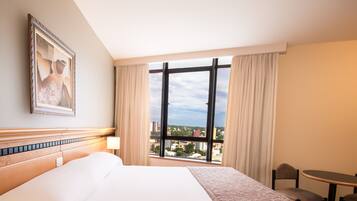 Quarto de hotel com uma cama grande, uma janela com cortinas, uma mesa pequena e um quadro na parede.