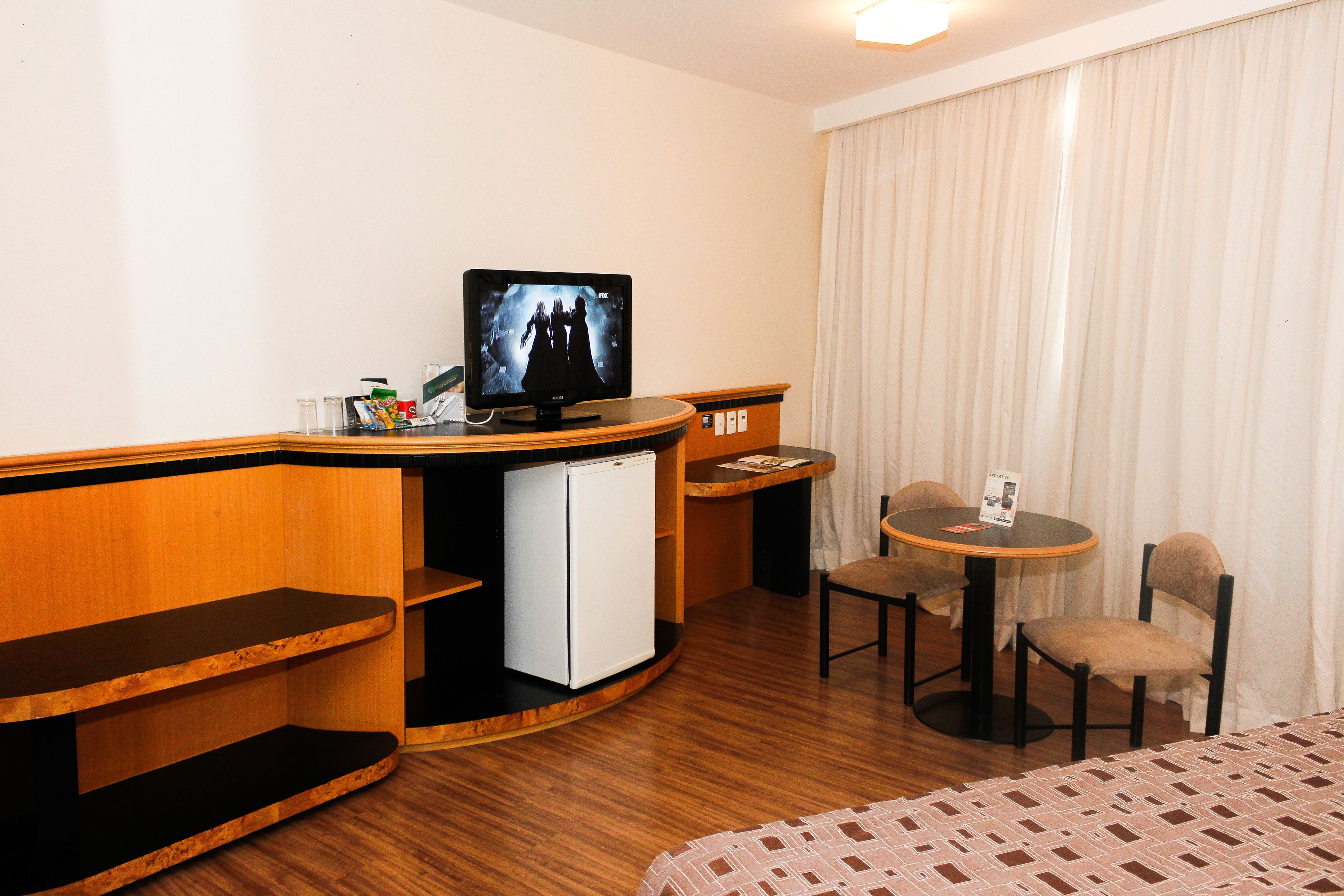 A hotel room with a wooden desk, a television, a mini-fridge, a small table with chairs, and a bed with a patterned bedspread.