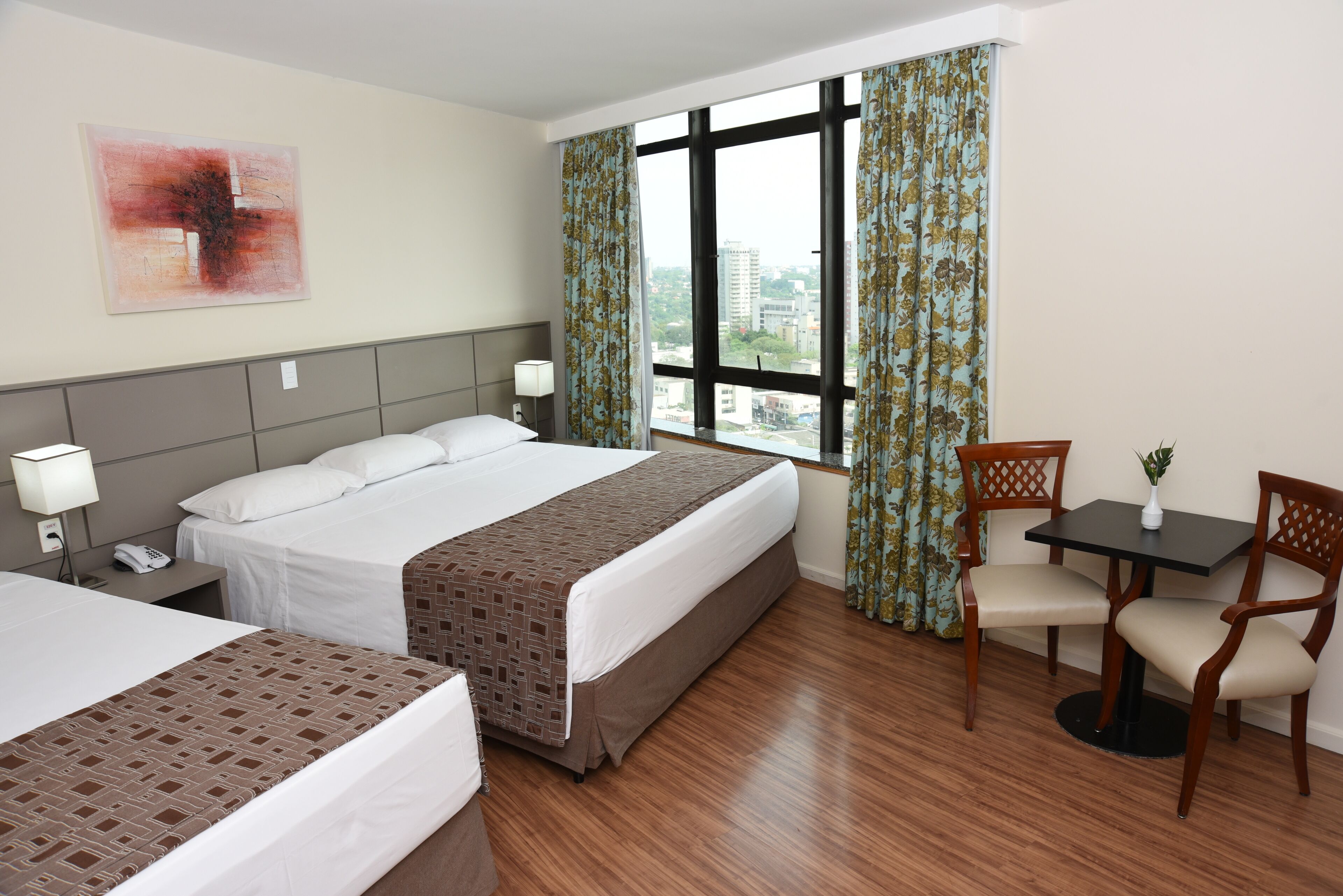 A hotel room with two beds, a desk with a chair, a window with curtains, and a painting on the wall.