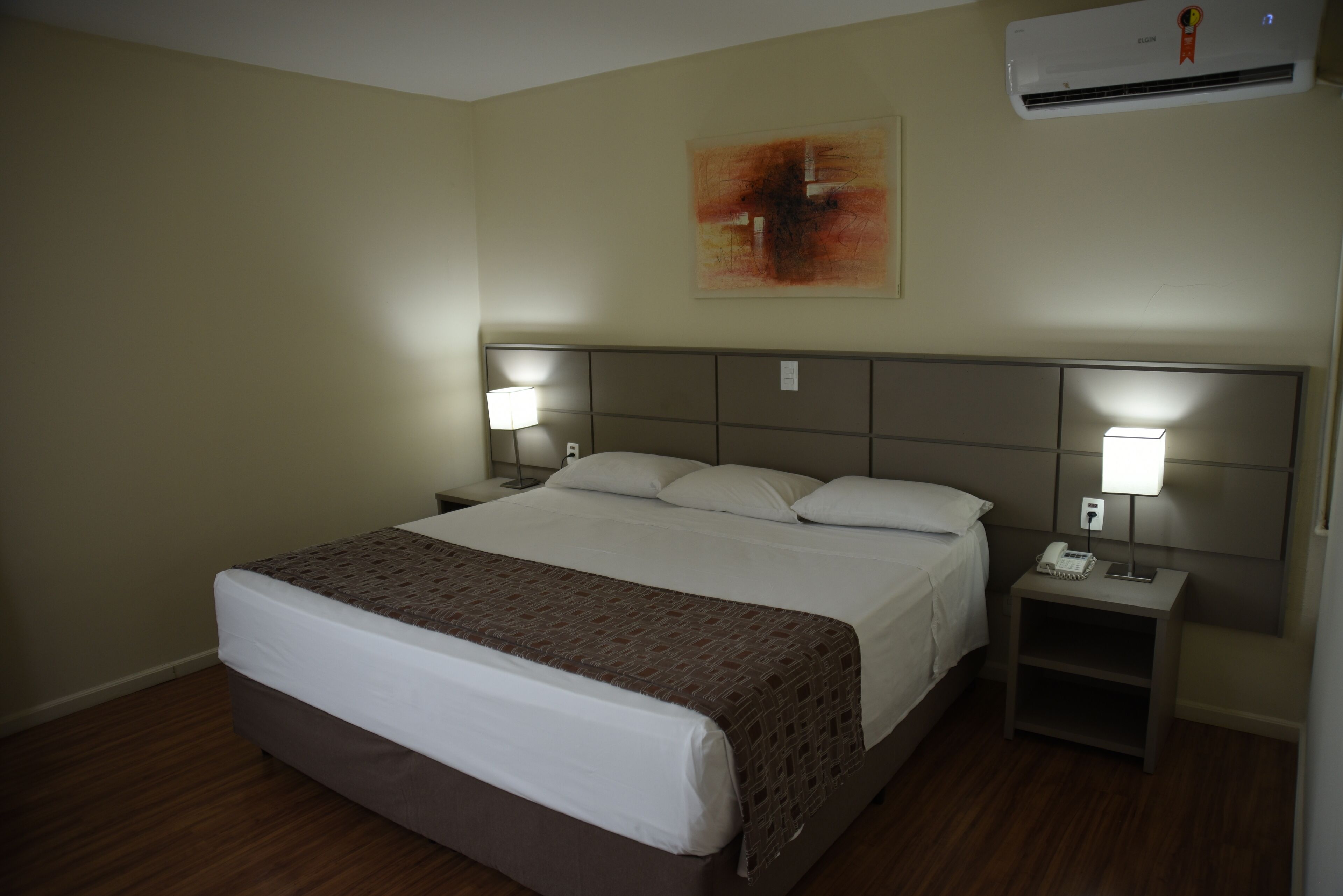 Une chambre d’hôtel avec un grand lit, deux tables de chevet avec des lampes, un climatiseur fixé au mur et un tableau au-dessus du lit.